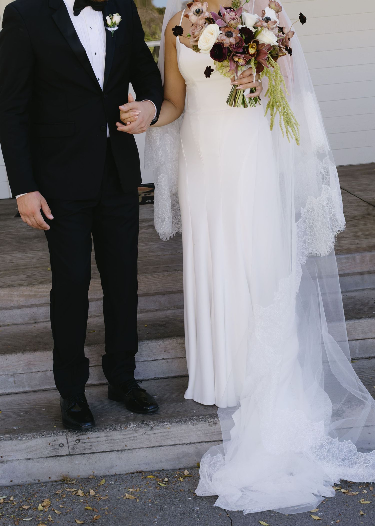 Wedding couple stands on white steps with elegant floral bouquet and formal attire.