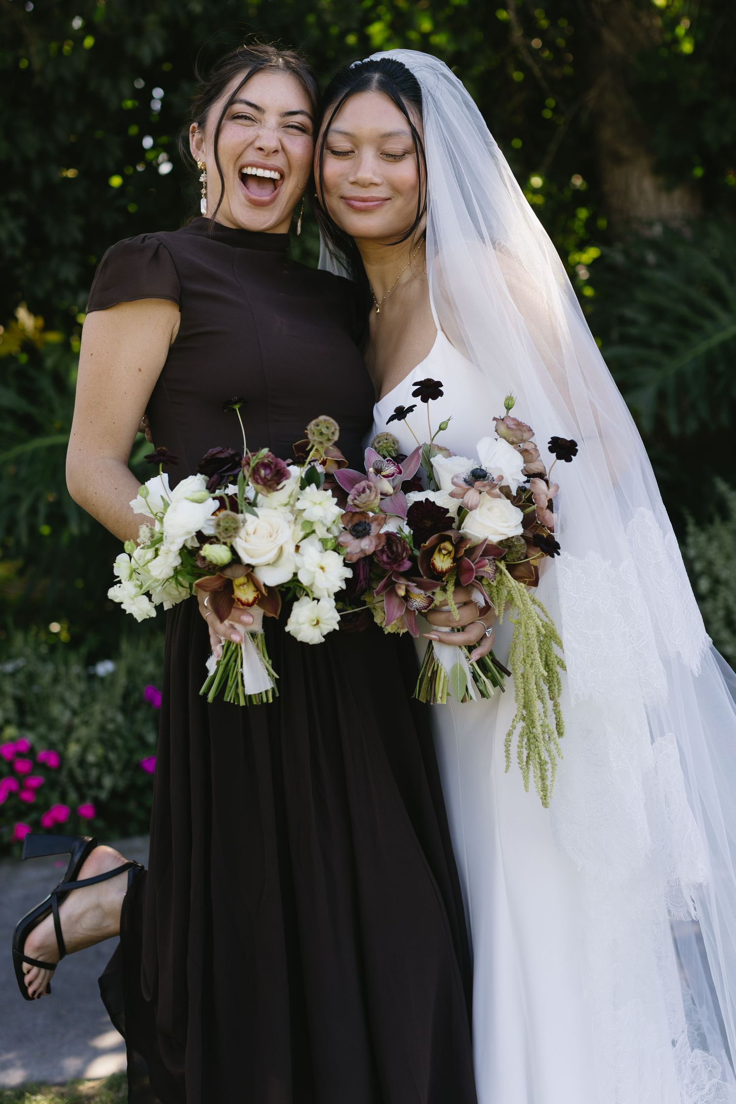Bride in white gown and bridesmaid in black dress hold matching floral bouquets while laughing together.