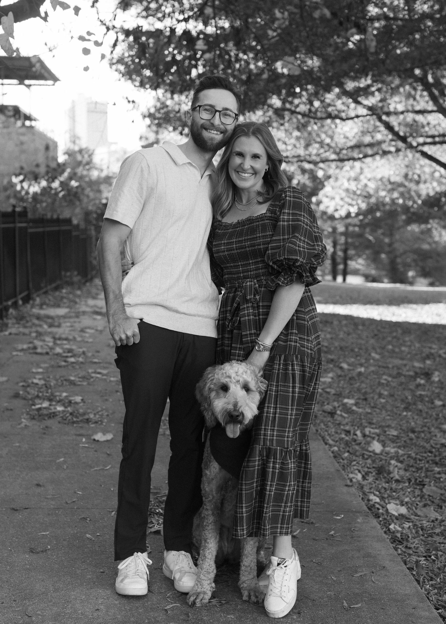 A couple and their goldendoodle pose for black and white photos on a leaf-covered path.
