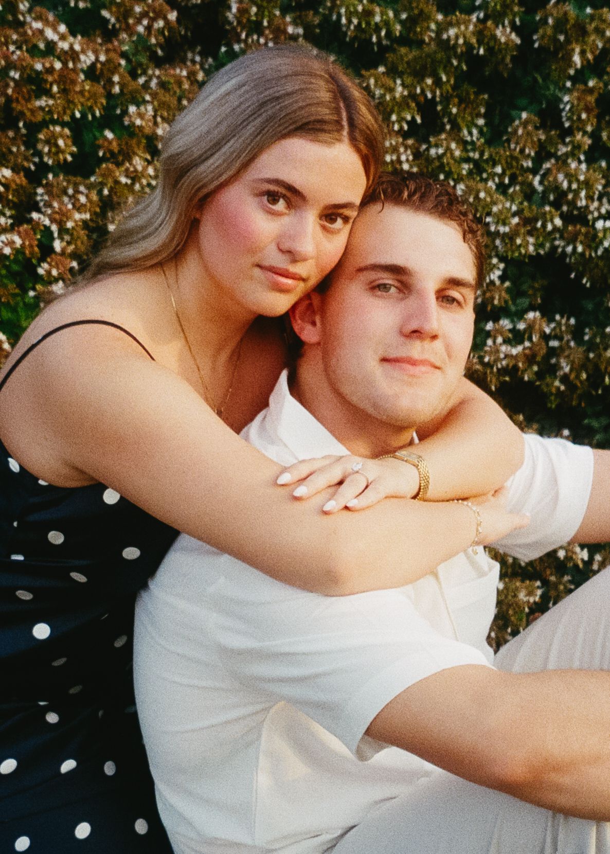 A couple embraces outdoors in casual attire with polka dot dress and white t-shirt against a sunlit floral background.