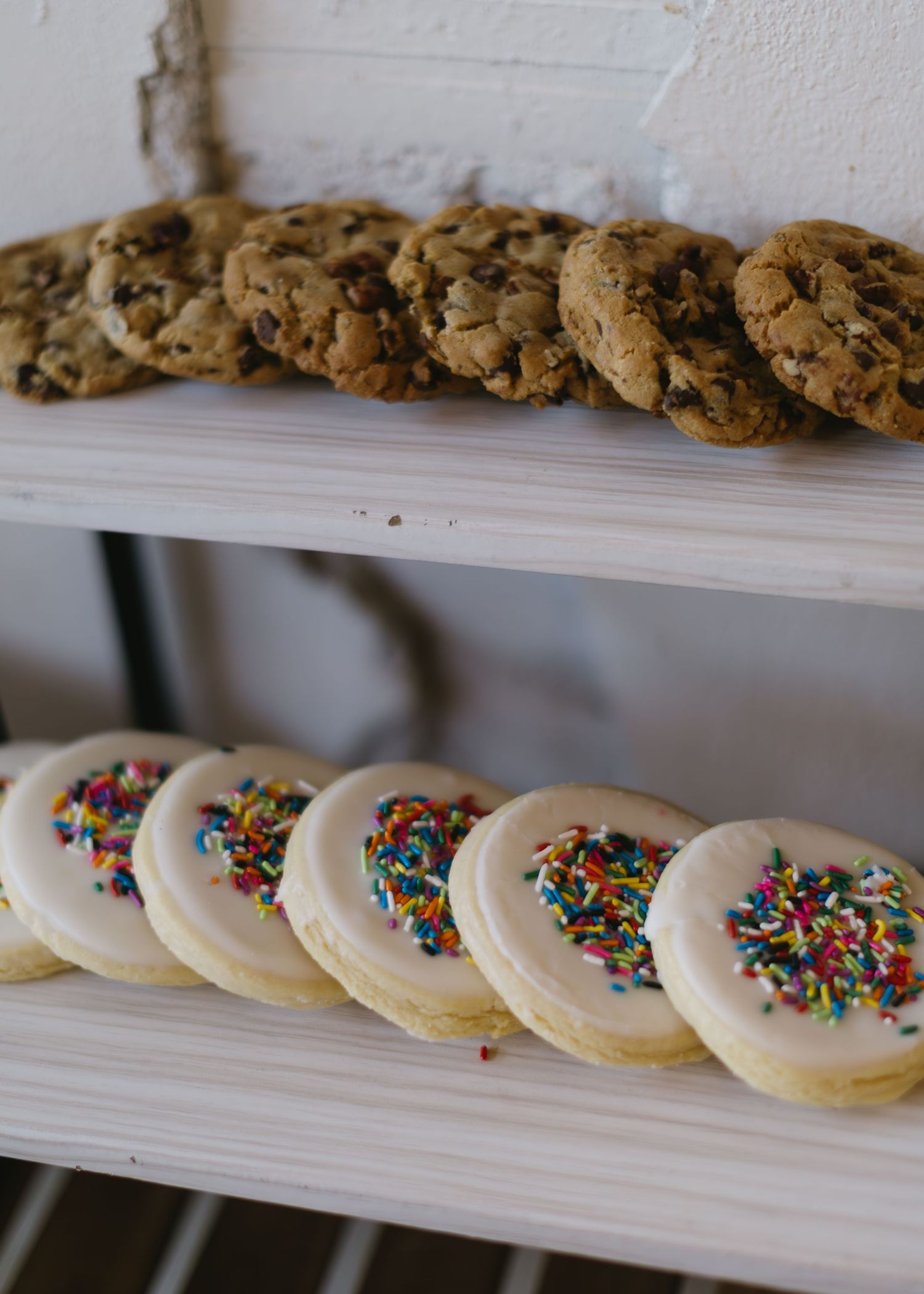 Rows of cookies arranged on white shelves, featuring chocolate chip and frosted sugar cookies with sprinkles.