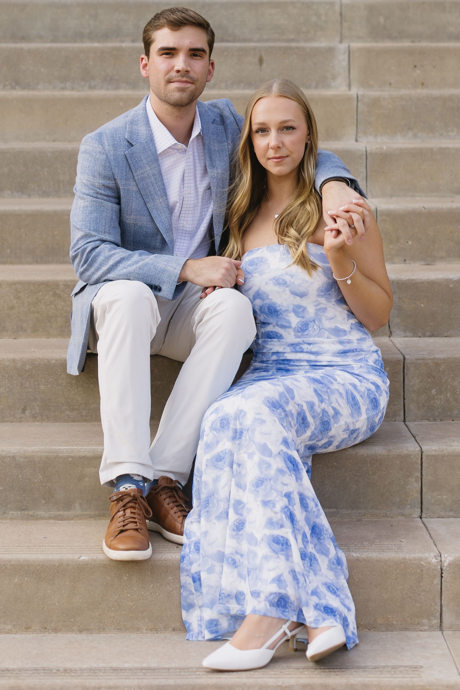 A couple in formal attire sits together on concrete steps with the person wearing a blue and white tie-dye dress.