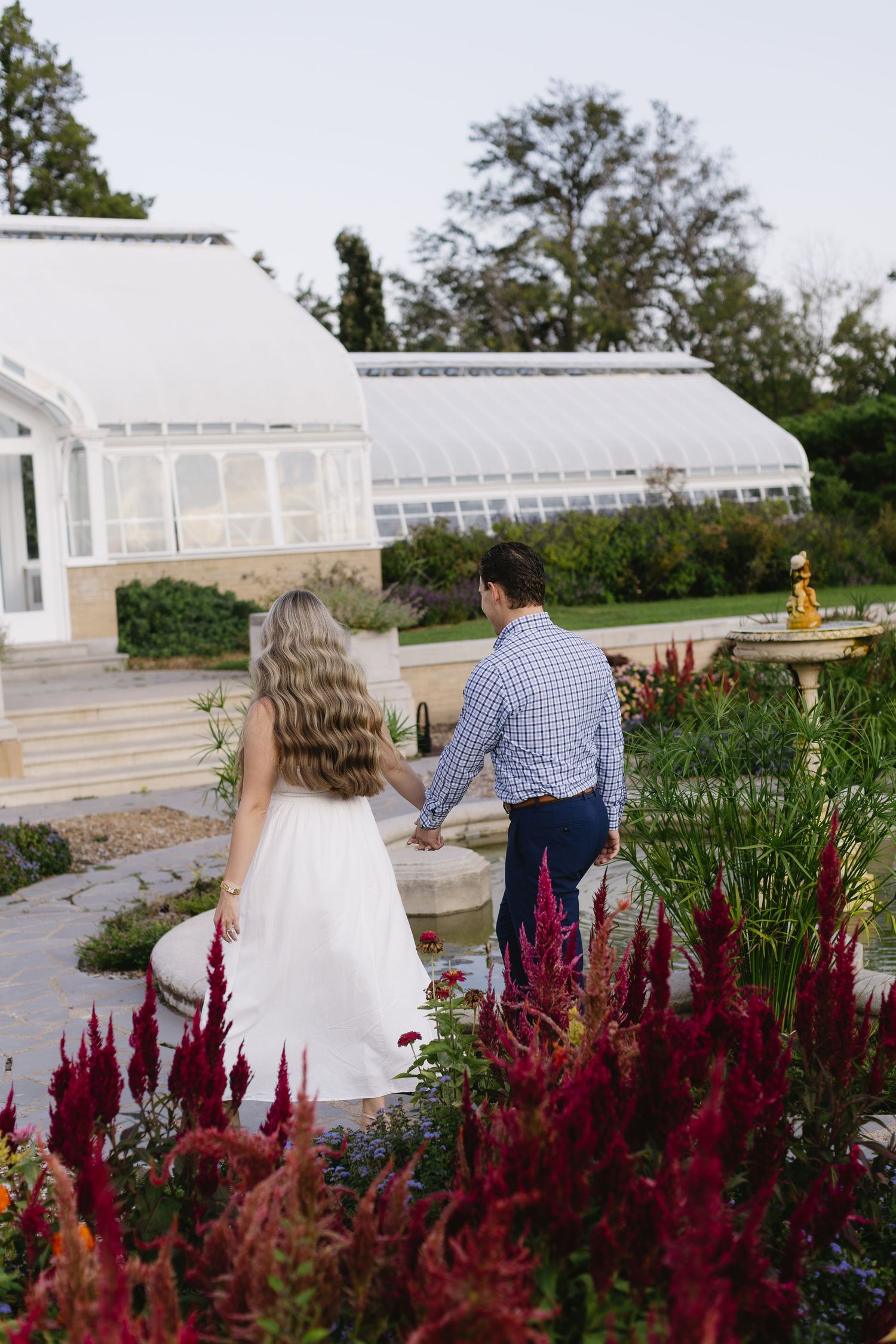 Couple walking hand in hand past vibrant red flowers near a greenhouse.