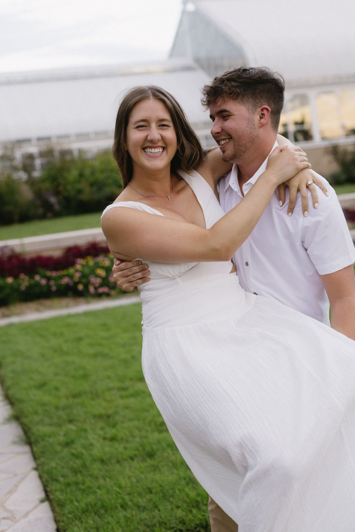 A joyful couple in white outfits share a candid moment as he lifts her on a manicured lawn.