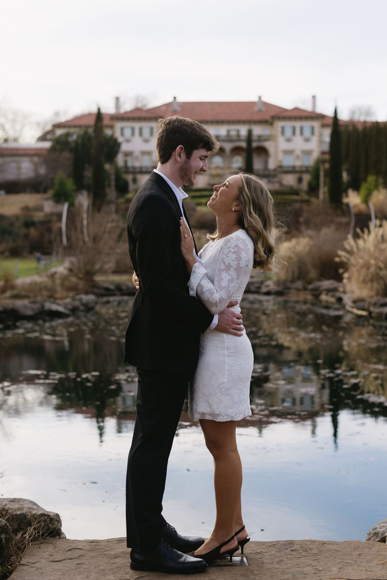 Couple in formal wear poses romantically by a reflective pond with buildings in background.