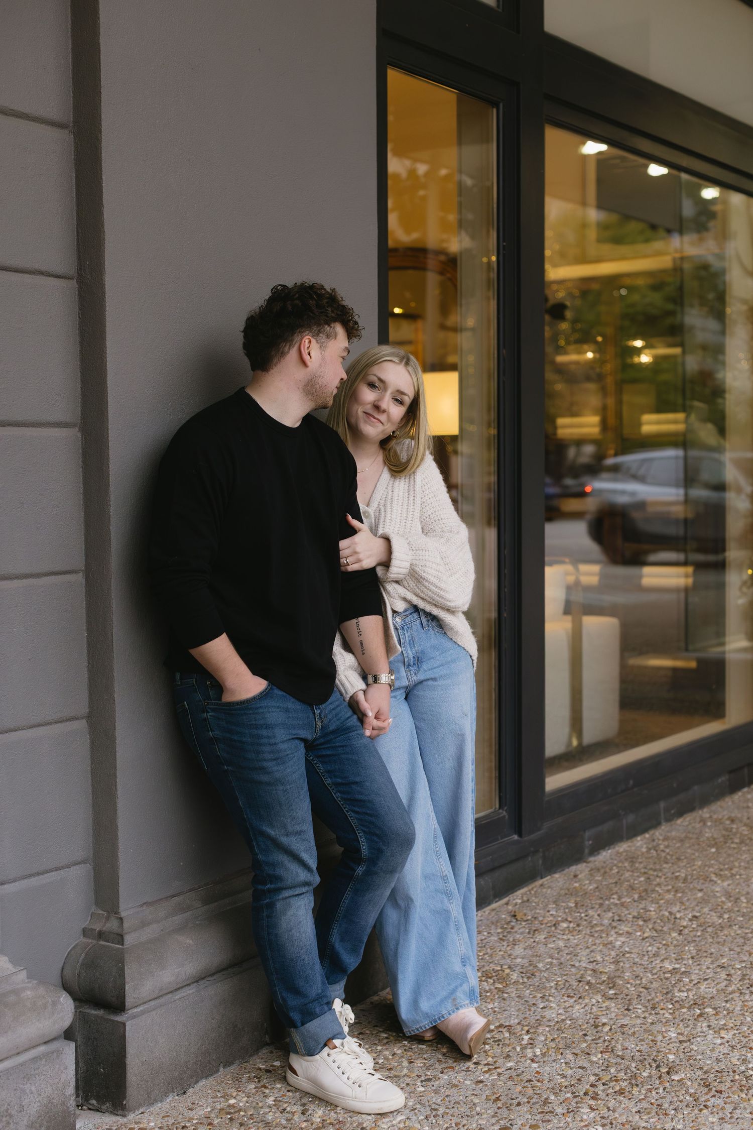 A couple in casual wear sharing playful moments against a storefront window.