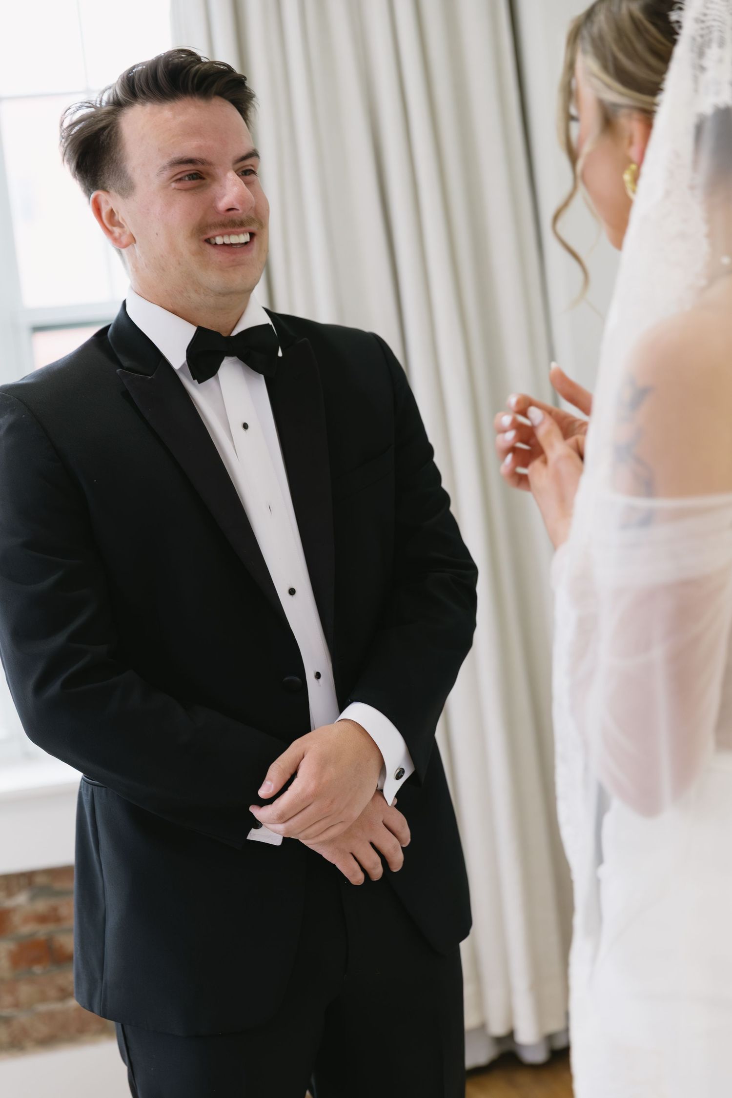Wedding couple sharing a joyful moment in formal attire against white curtains.