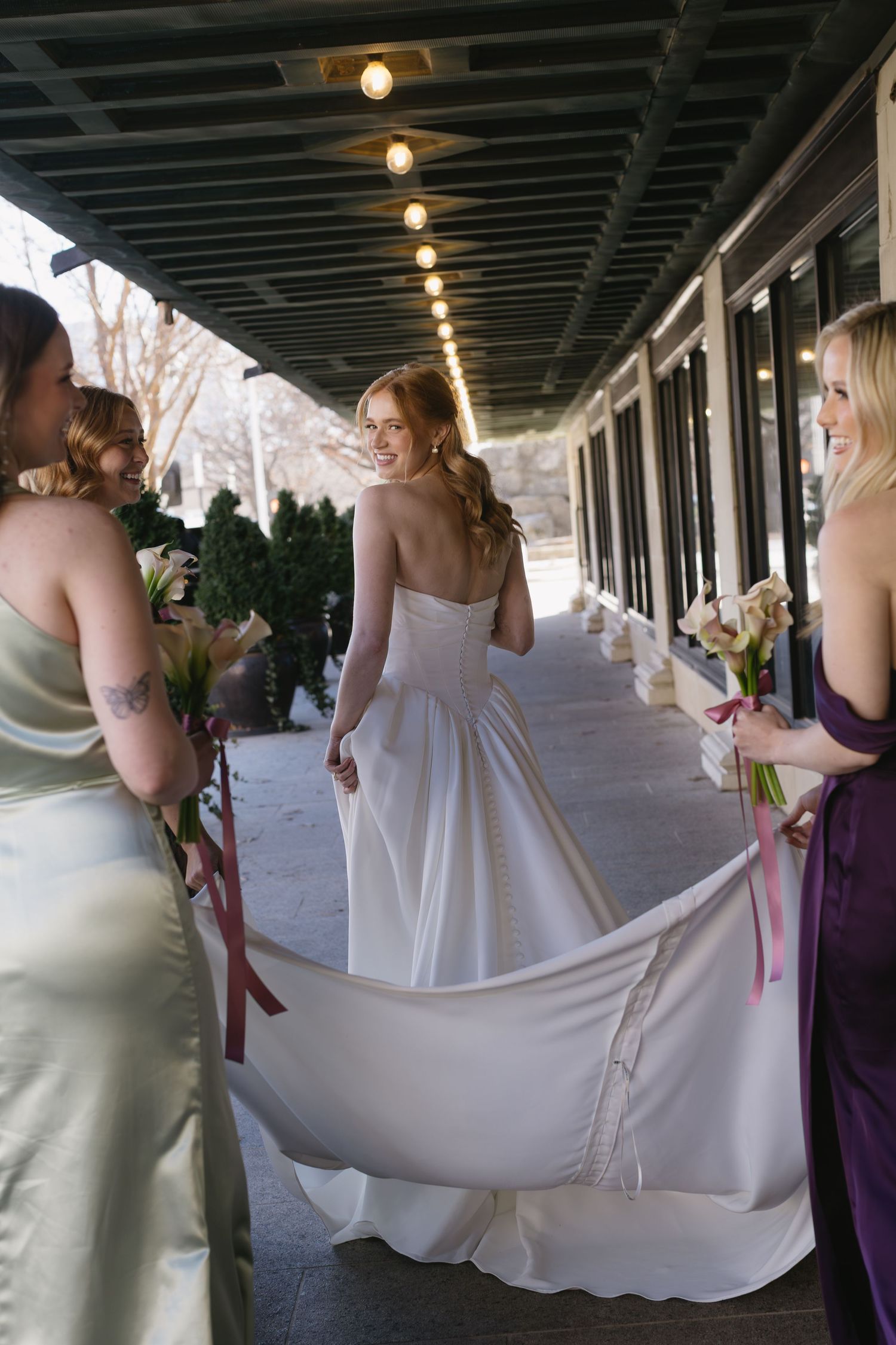 Bridesmaids in purple dresses help arrange a bride's flowing white wedding dress train in an outdoor corridor.