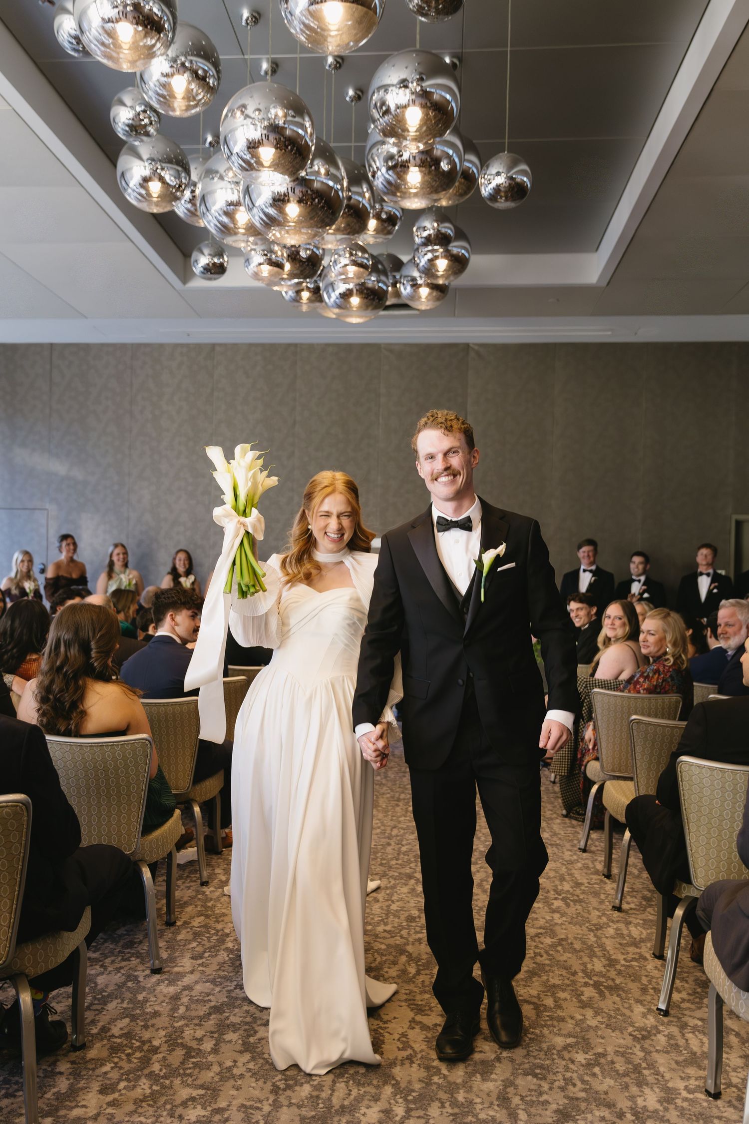 Couple exits wedding ceremony under modern chrome bubble chandelier as guests look on.