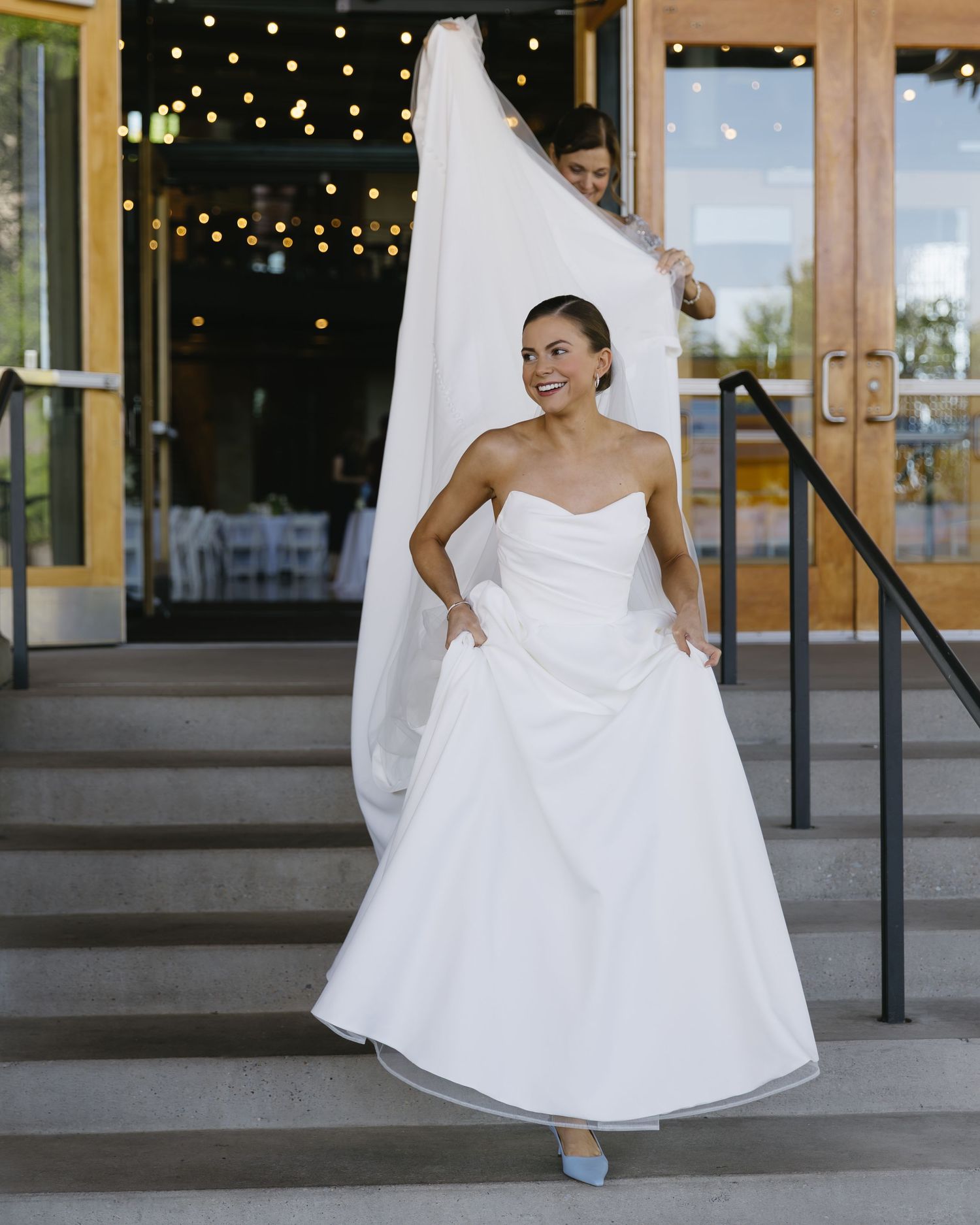 A bride in a classic white strapless ball gown poses on stone steps with string lights in the background.