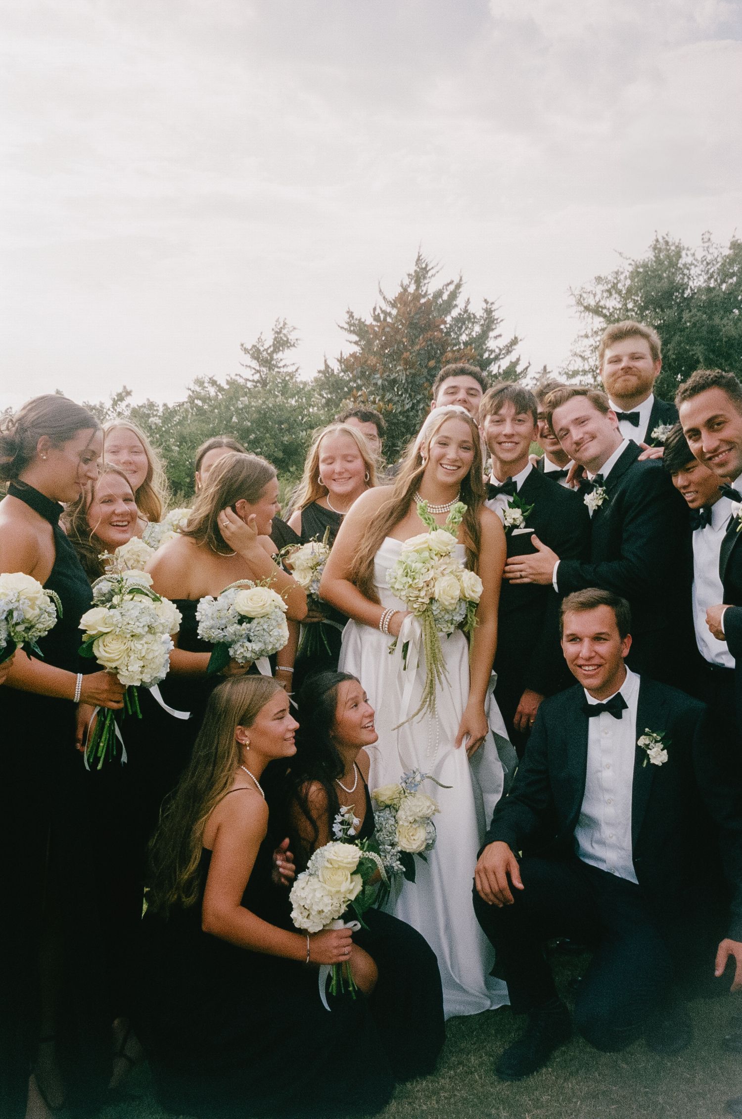 Wedding party in formal black and white attire poses together outdoors while laughing and celebrating.
