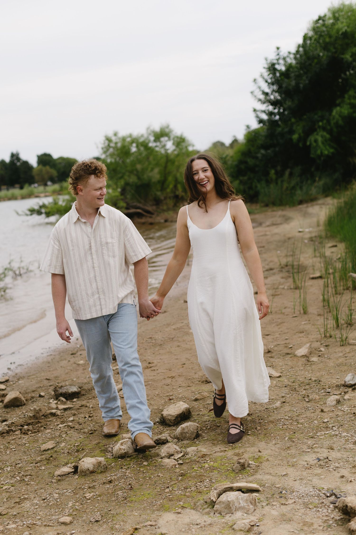 A couple walks hand in hand along a rocky shoreline on a summer day.