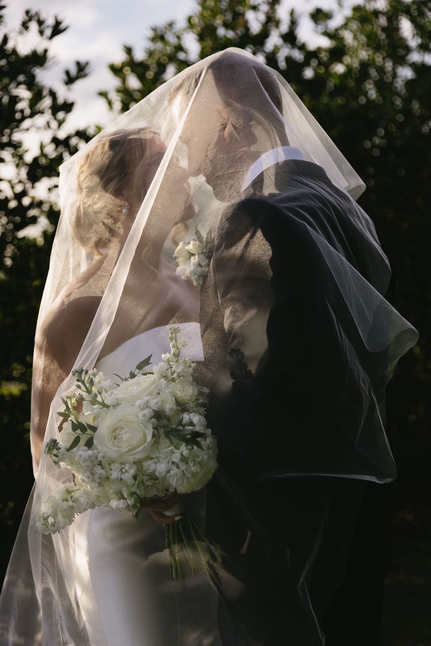 Bride and groom share a romantic moment under a flowing veil with white floral bouquet during an outdoor wedding ceremony.