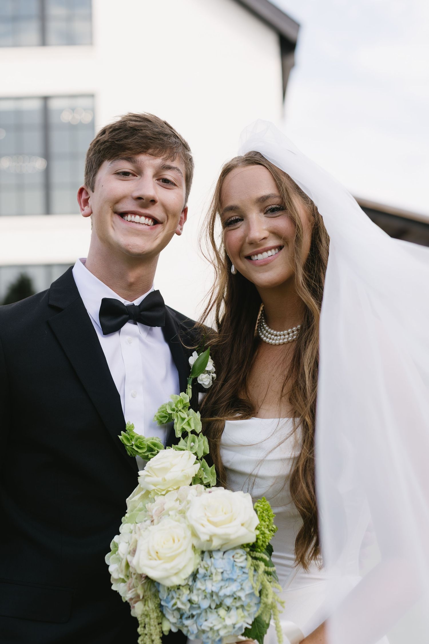 Newlyweds smiling together while holding a bouquet of white roses and blue hydrangeas.