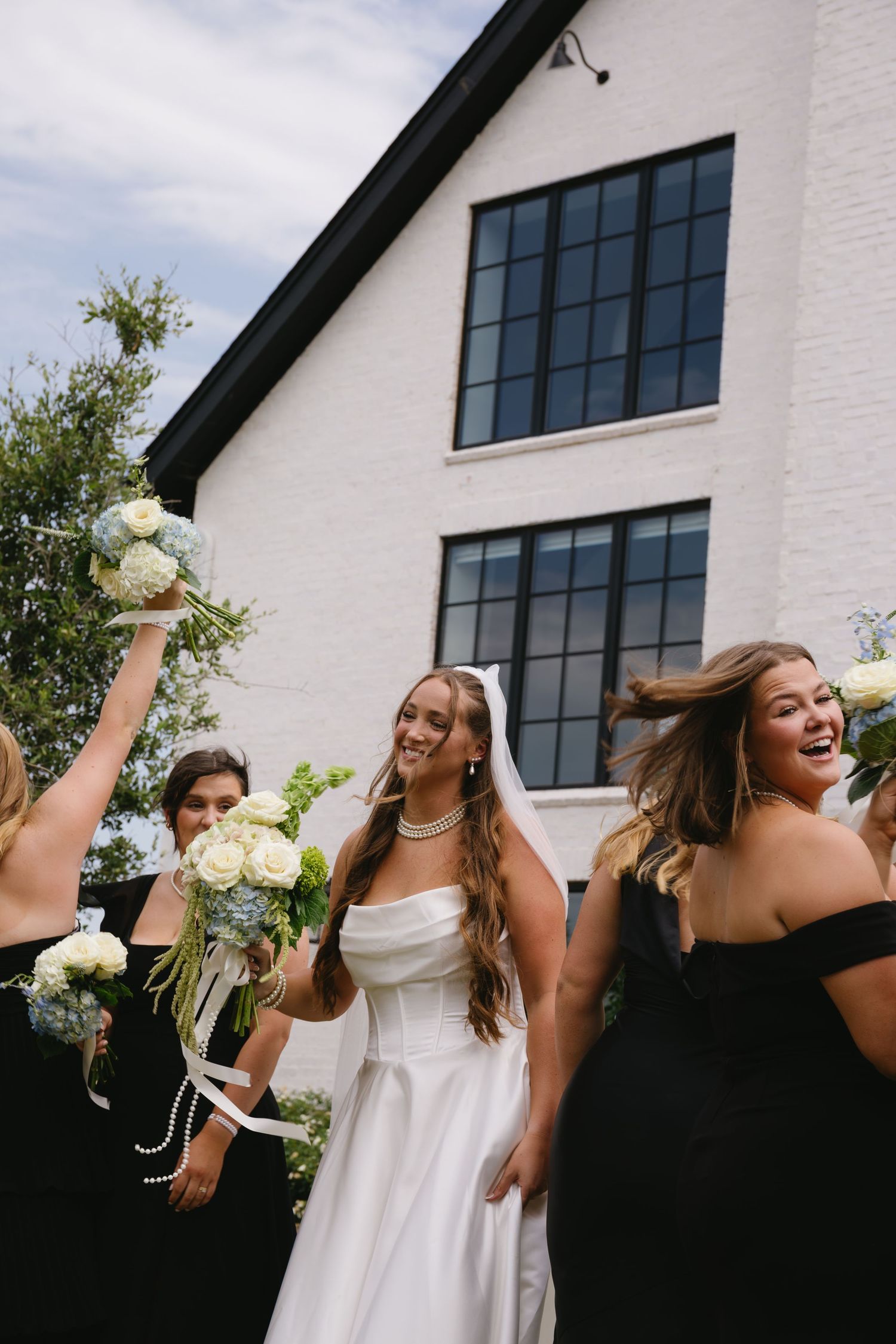 Bridesmaids in black dresses playfully pose outdoors against a white modern building holding white bouquets.