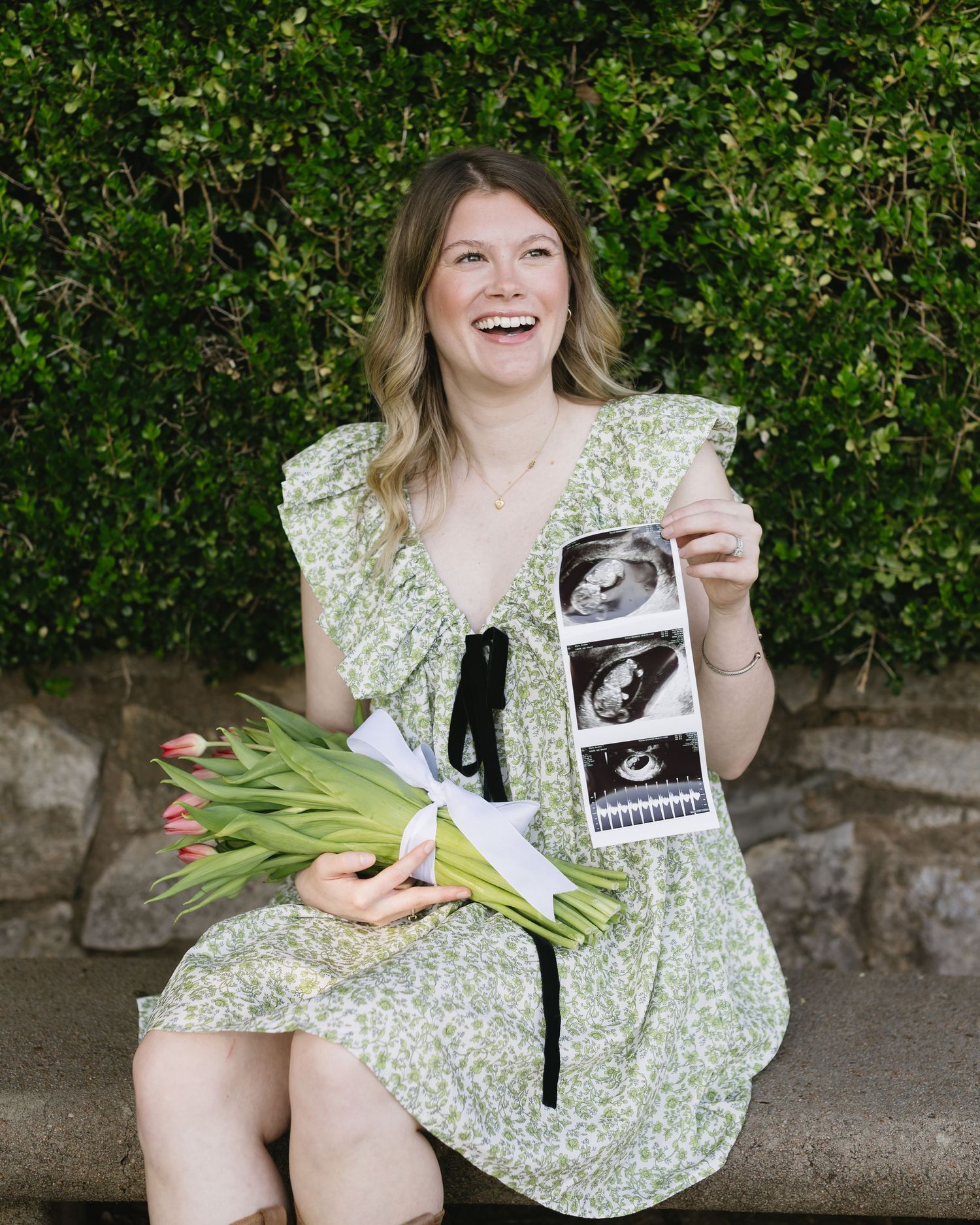 A woman in a floral dress holds tulips and sonogram photos while sitting outdoors.