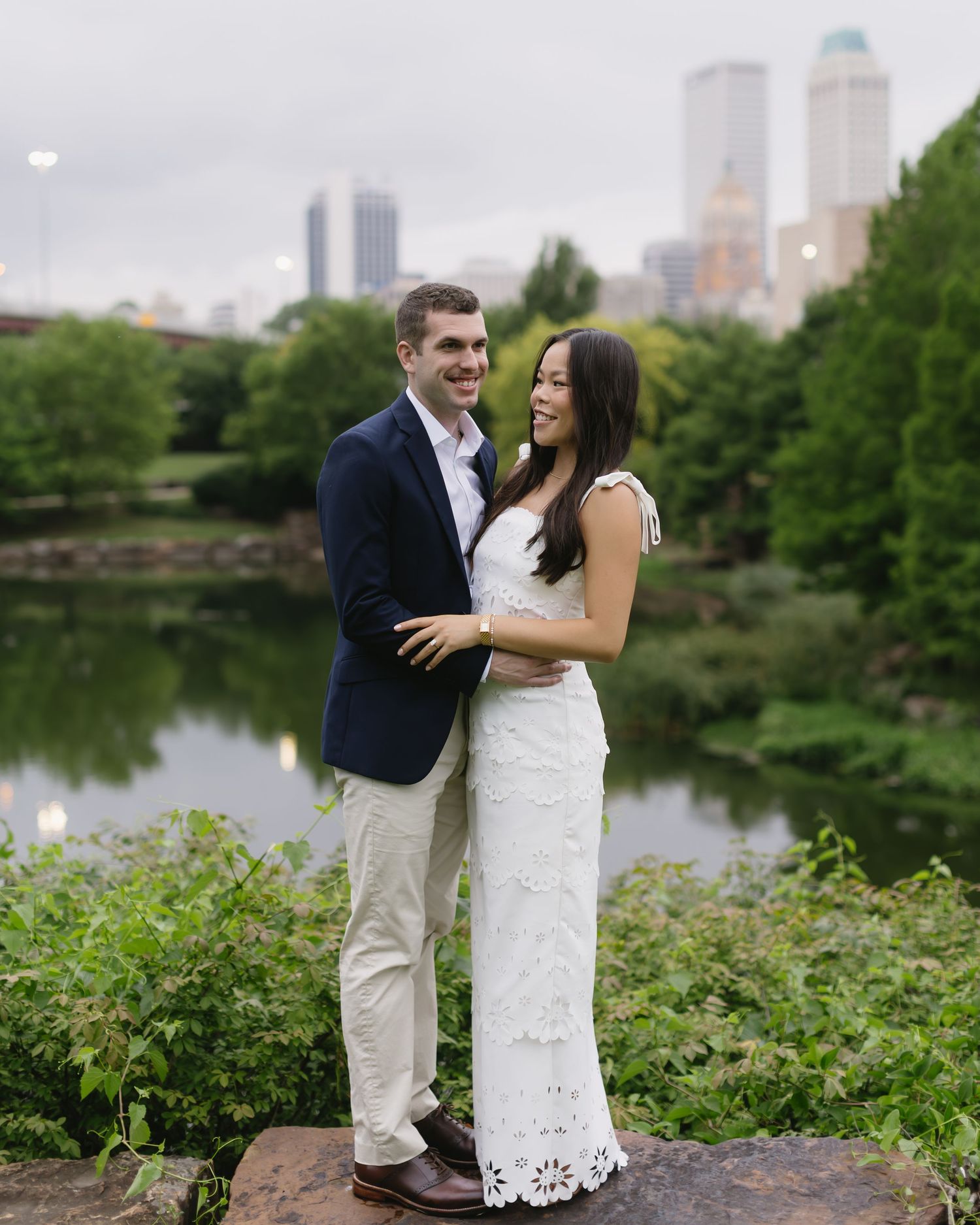 Romantic couple poses by lake with urban skyline and trees in background.