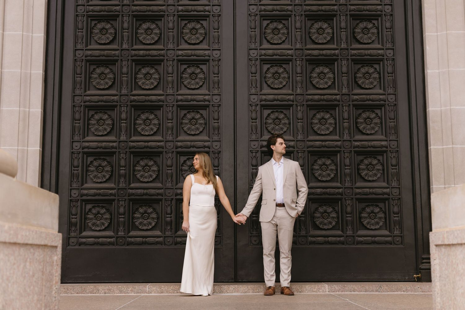 A couple in formal attire poses against large decorative doors of a historic building.