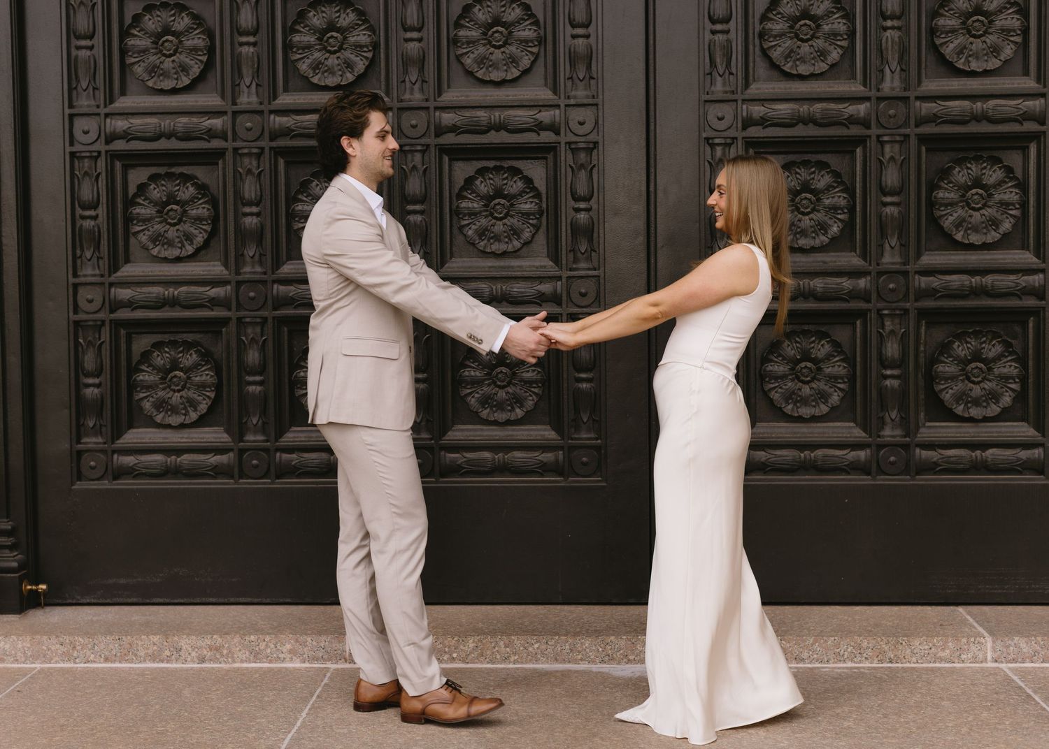 A couple in light-colored formal wear poses playfully against dark ornate doors.