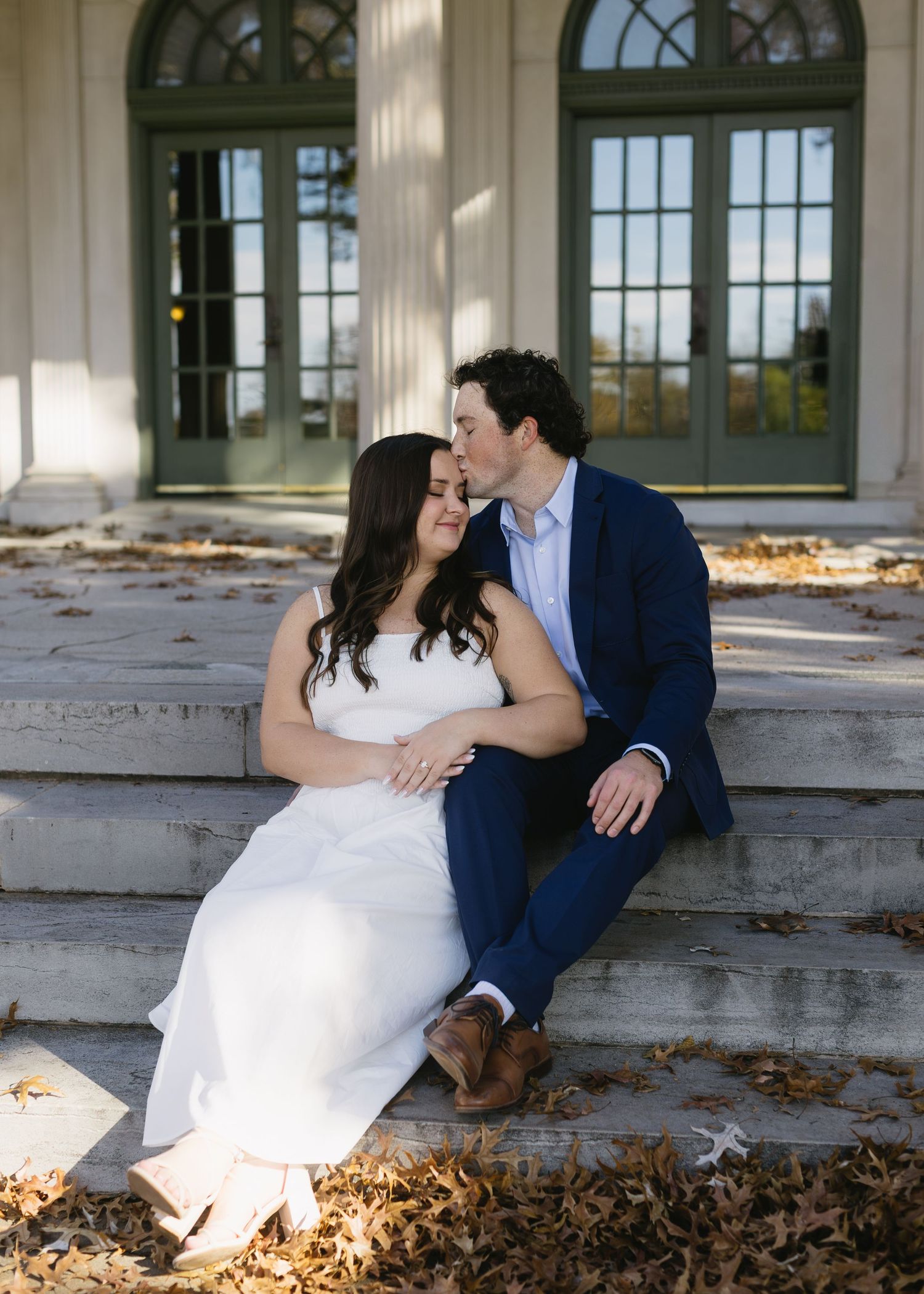 A couple sits on stone steps in formal attire with autumn leaves scattered around an elegant building entrance.