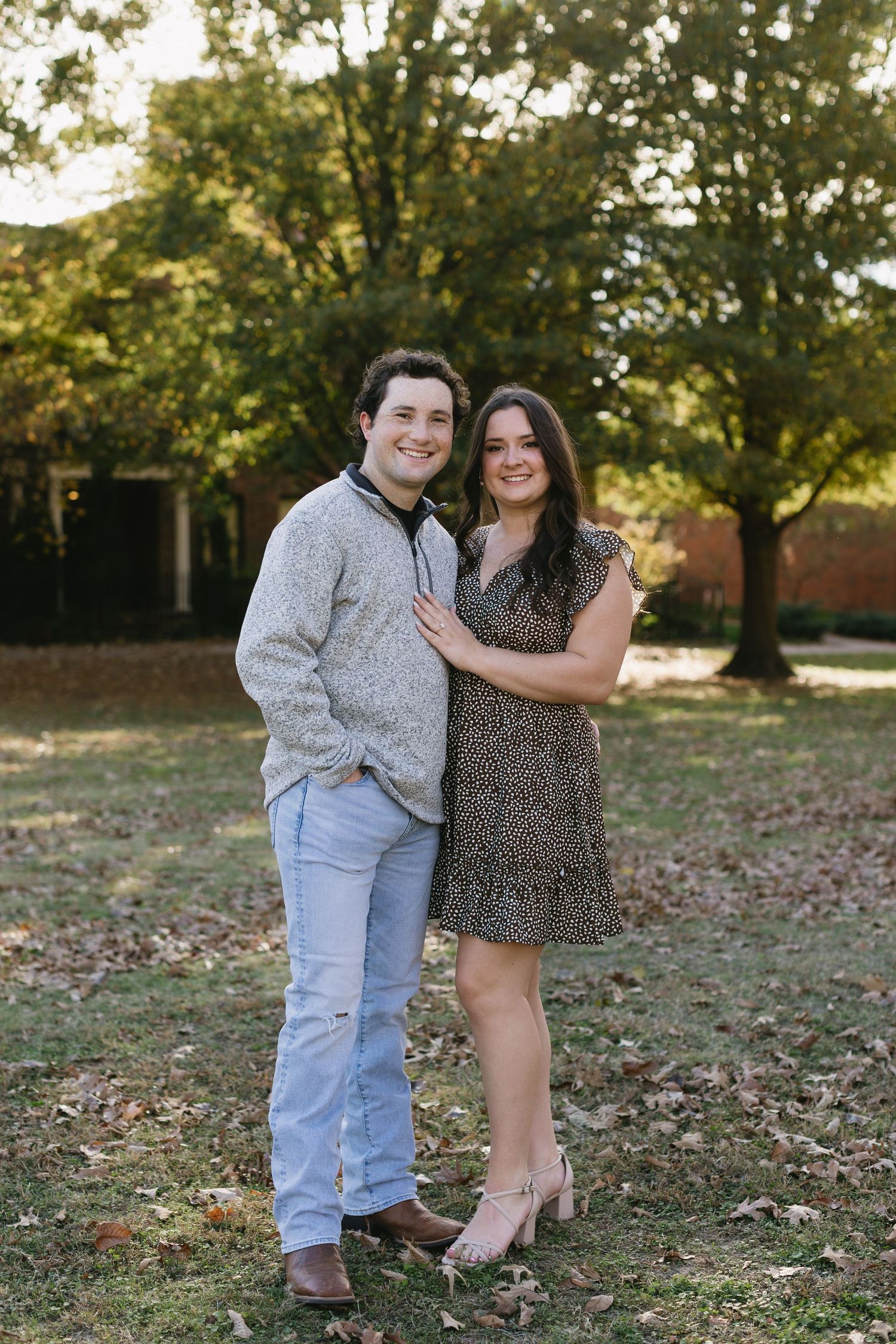 A couple poses playfully together on grass with autumn leaves and trees in the background.