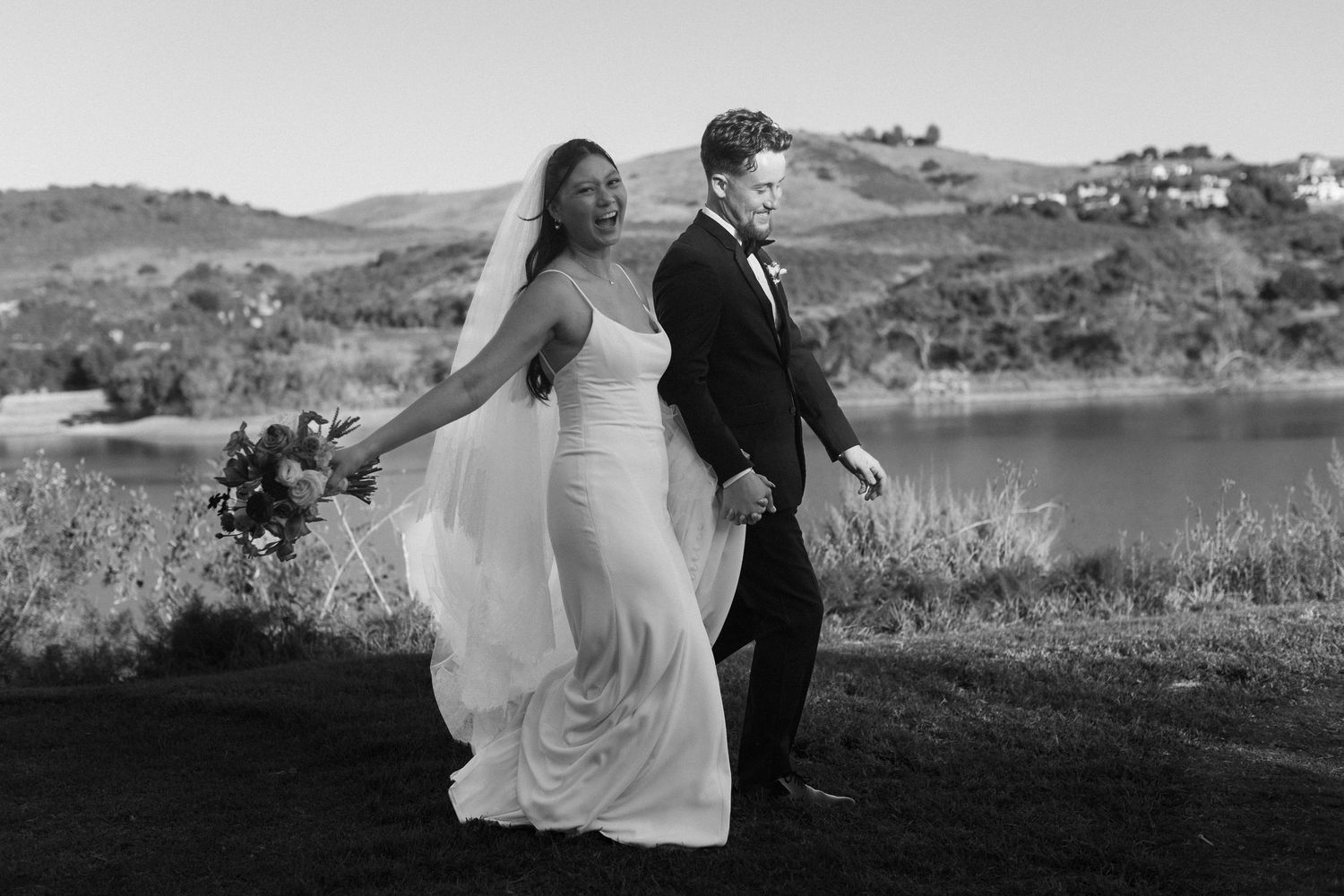 A couple in wedding attire dances together on a hilltop overlooking a scenic mountain landscape at sunset.