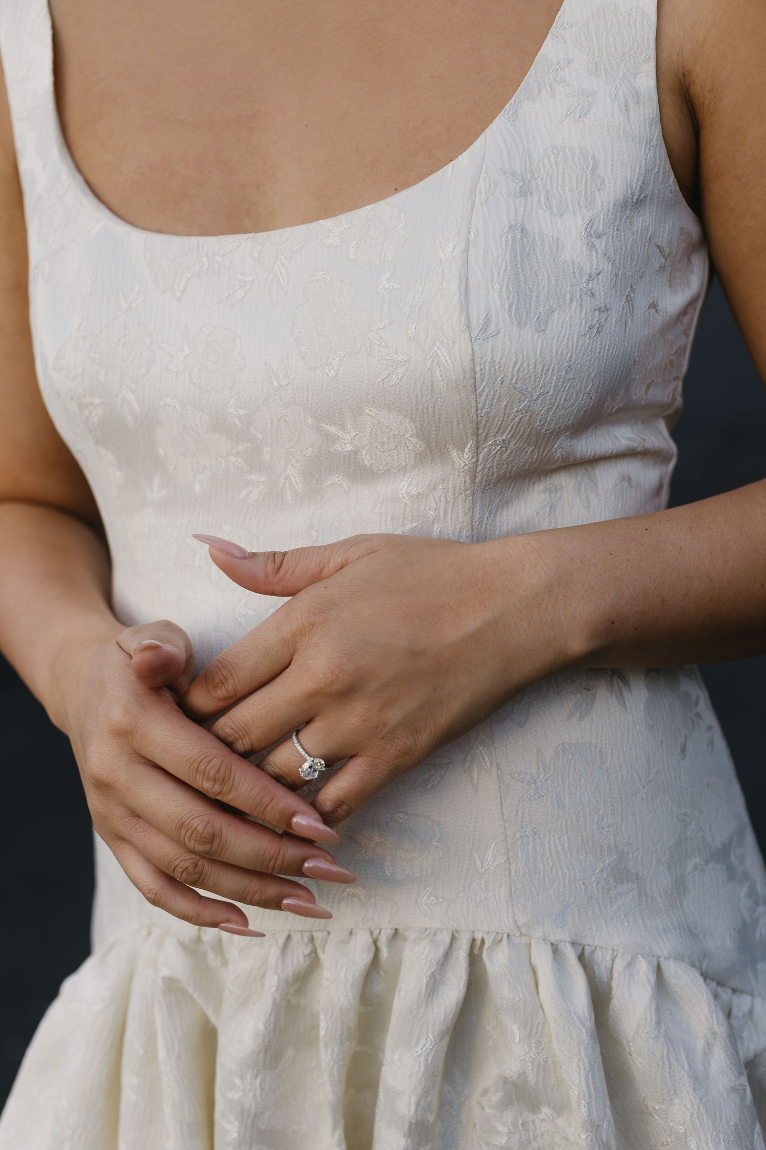 Close-up of hands with engagement ring on white lace dress with delicate floral pattern.
