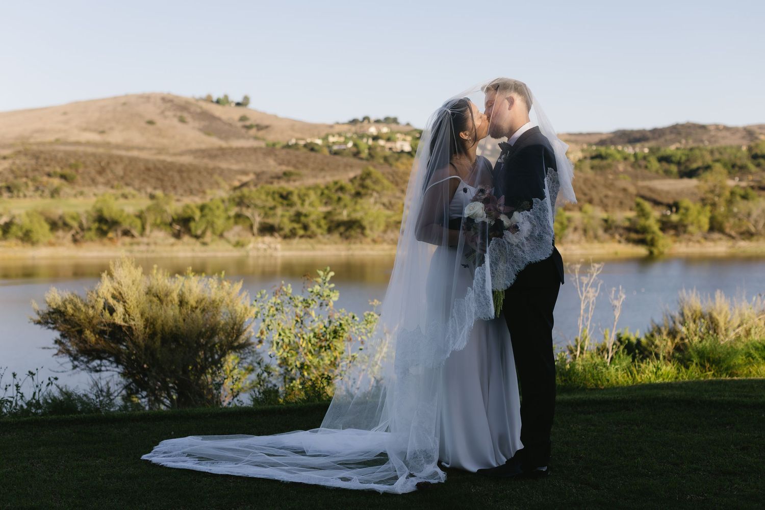 Newlyweds walking together against scenic lakeside landscape at sunset.