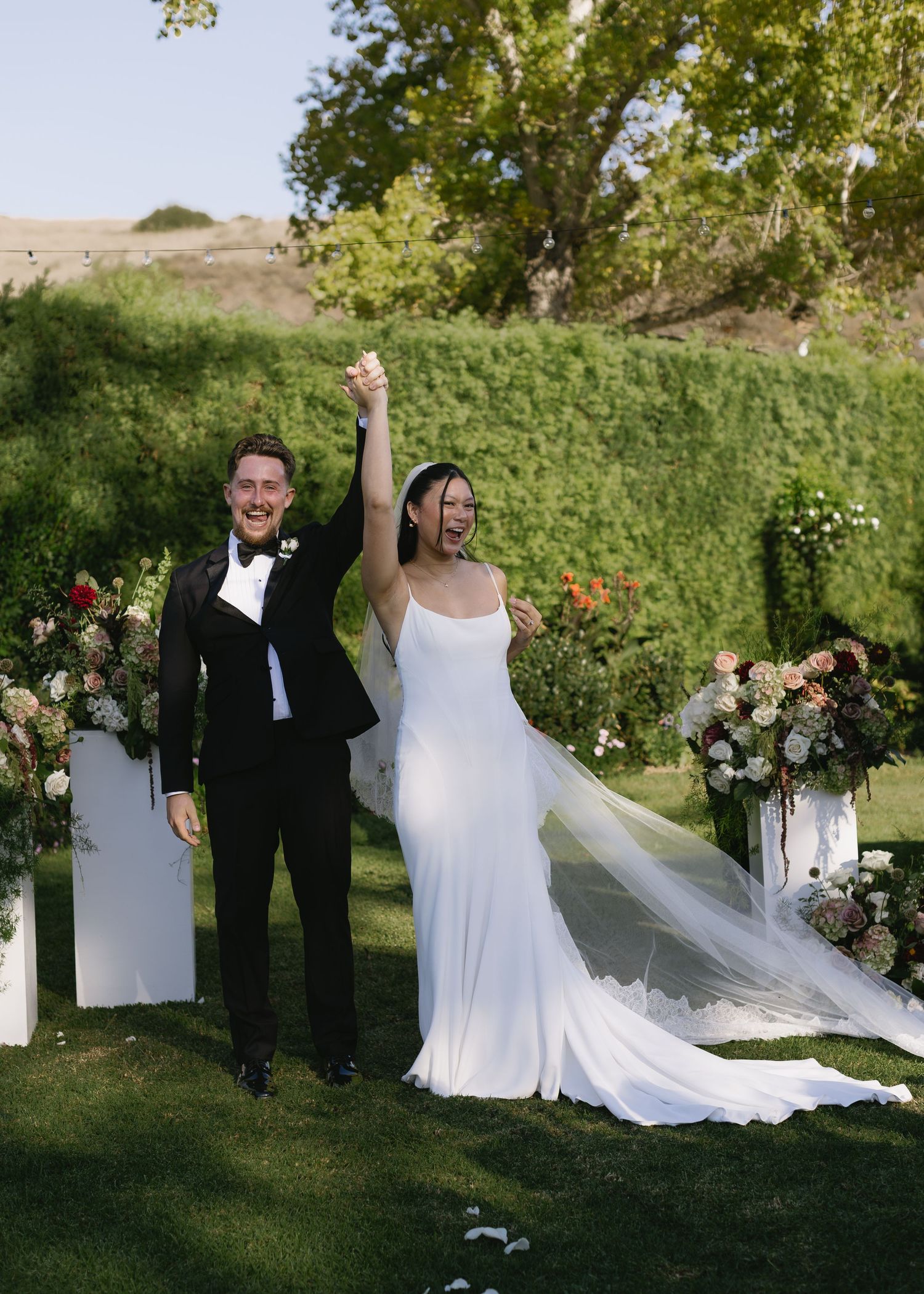 Elegant outdoor wedding ceremony with floral arch and flowing white dress against green landscape.