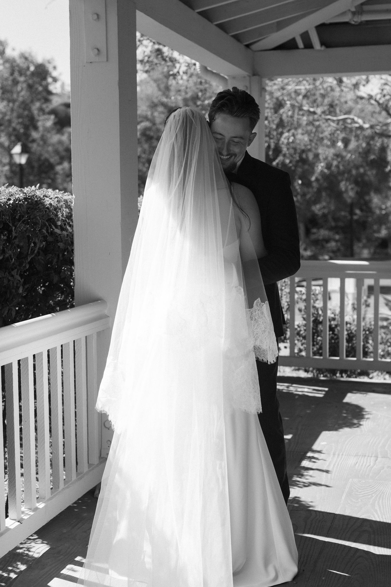 Wedding couple embracing on a sunlit porch with white columns and a flowing bridal veil.