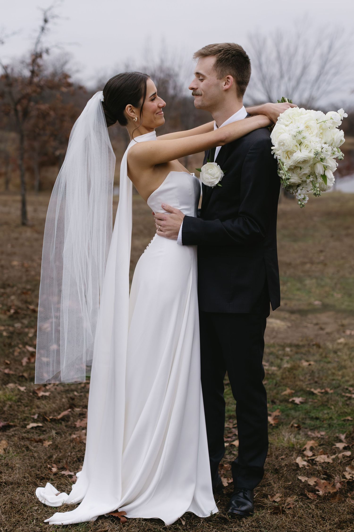 Couple embraces in romantic pose with white floral bouquet against autumn background.