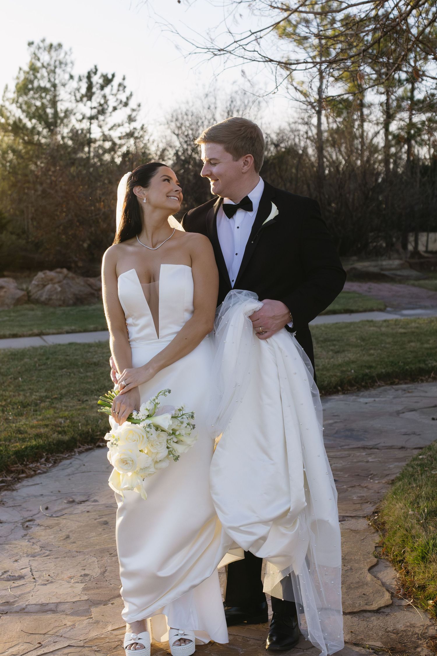 A couple in wedding attire dances and poses together on a stone pathway at sunset.
