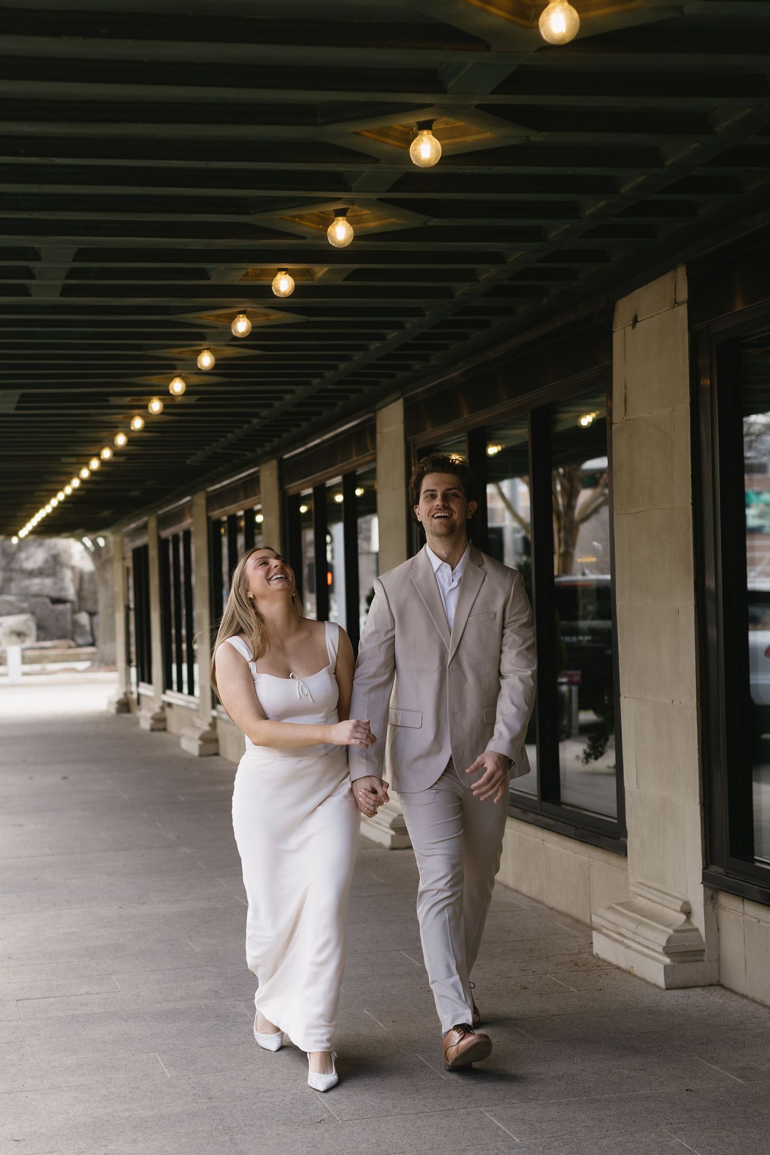 A couple strolls hand in hand under a covered walkway with string lights overhead.