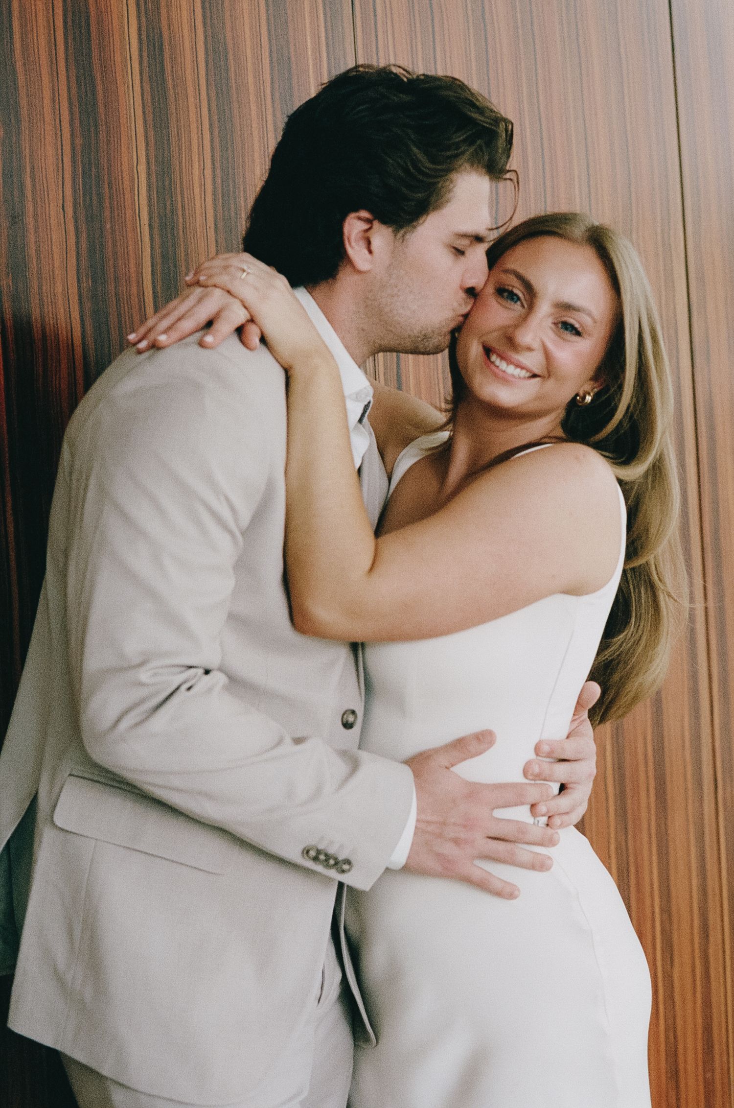 Tender moment between couple in white and beige outfits against wooden wall backdrop.