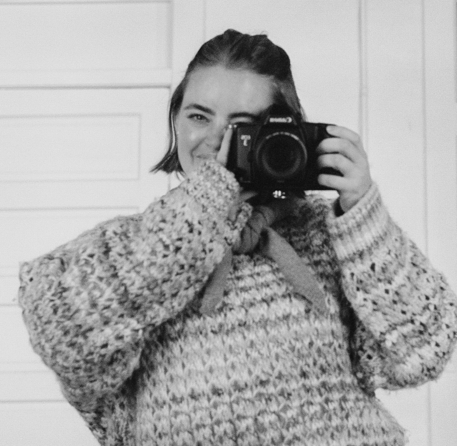 A person wearing a floral patterned sweater takes a self portrait in a mirror with a digital camera.