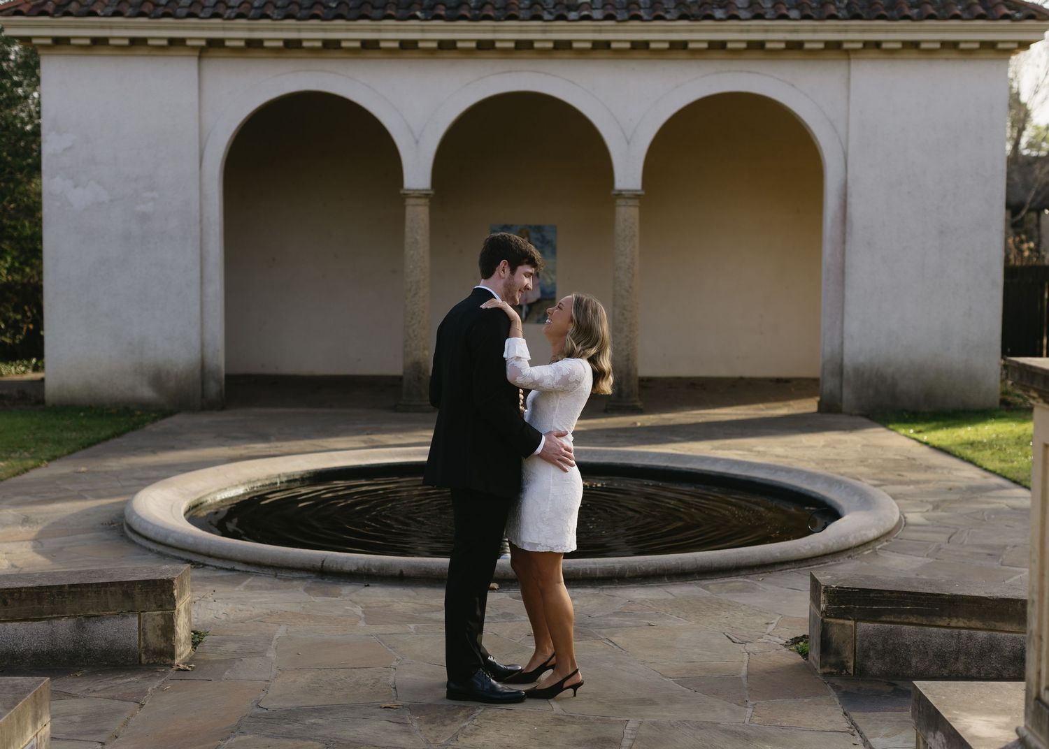 A couple in formal wear stands together by a fountain under arched columns.