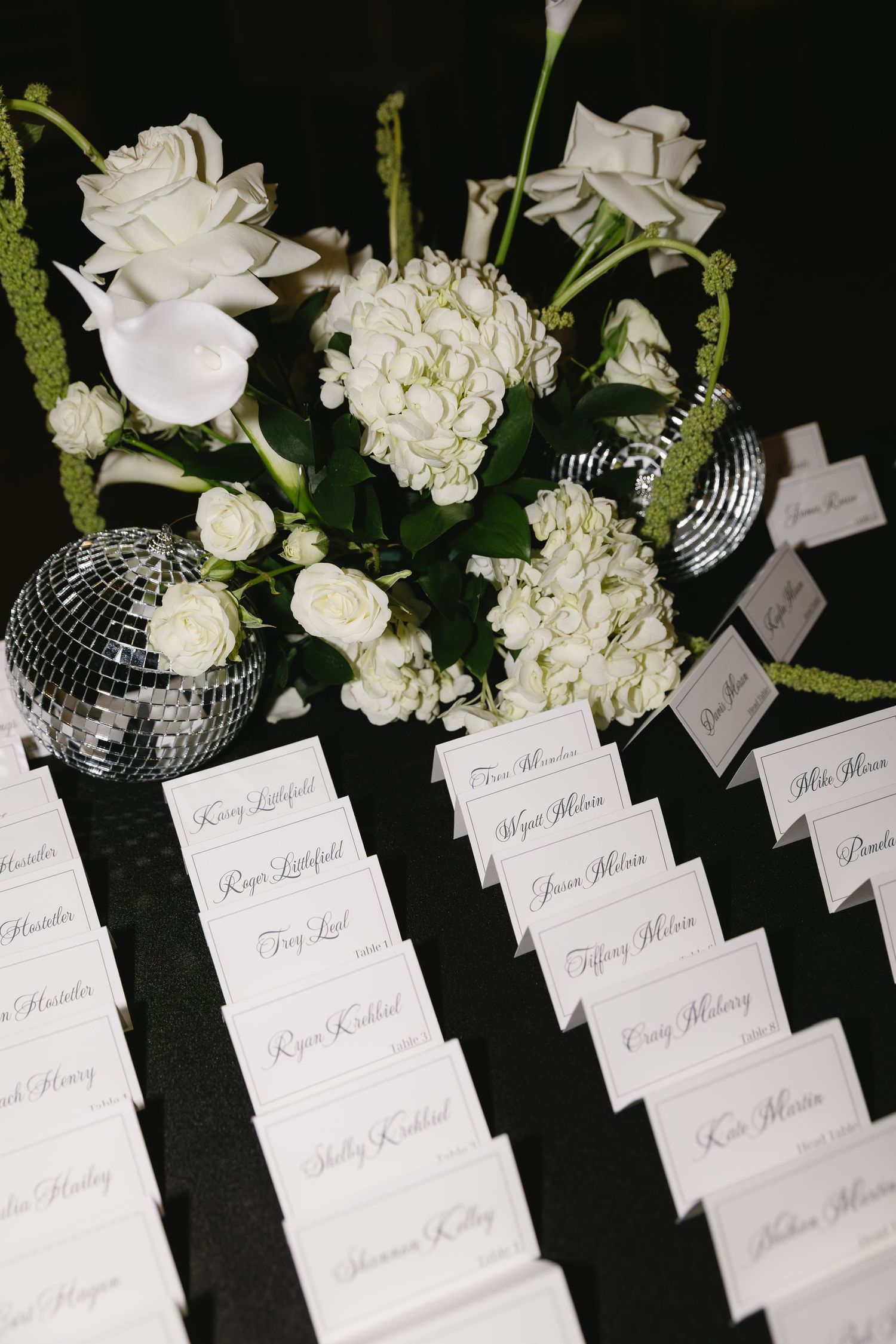 Elegant place cards with handwritten names displayed alongside white floral arrangements and mirrored disco balls.