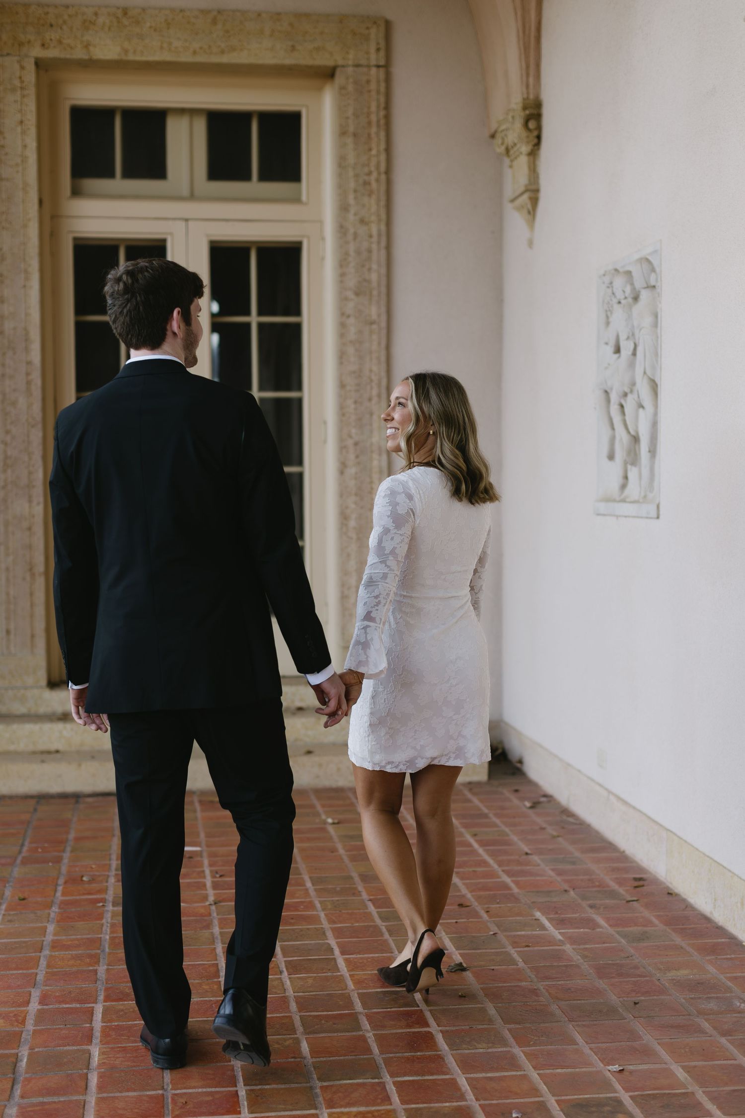 A couple in formal attire walks hand in hand along a covered walkway with terracotta tiles and classical architecture.