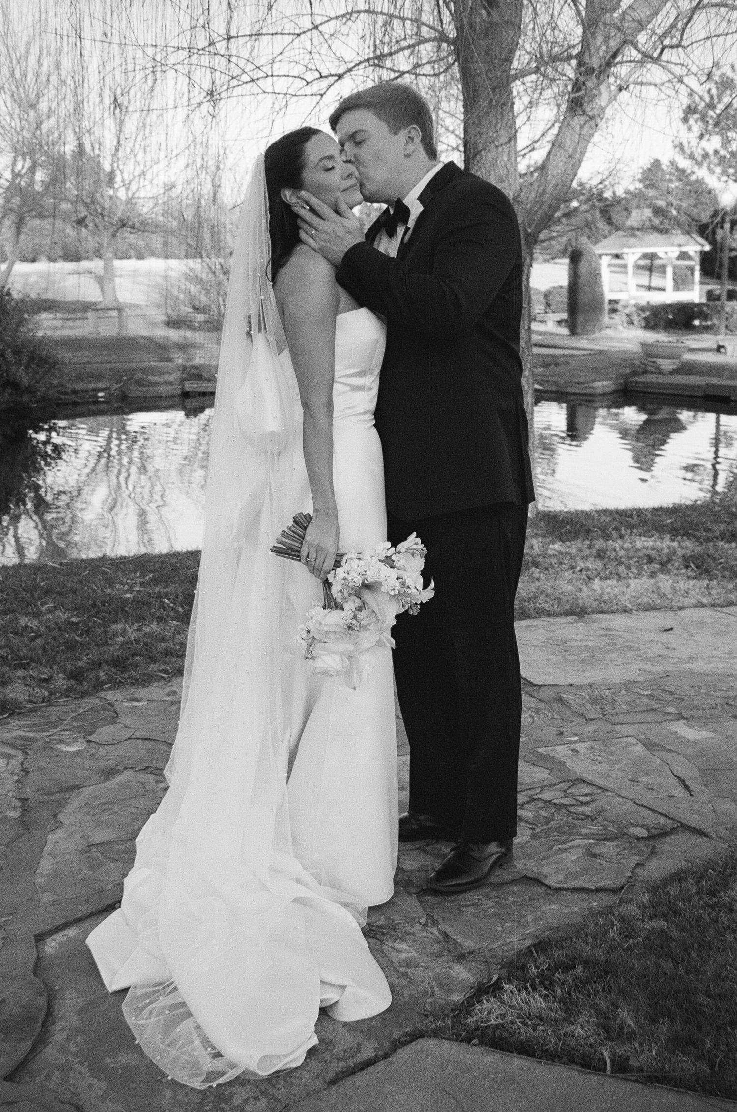 Newlyweds share romantic moment outdoors near stone pathway and winter trees.
