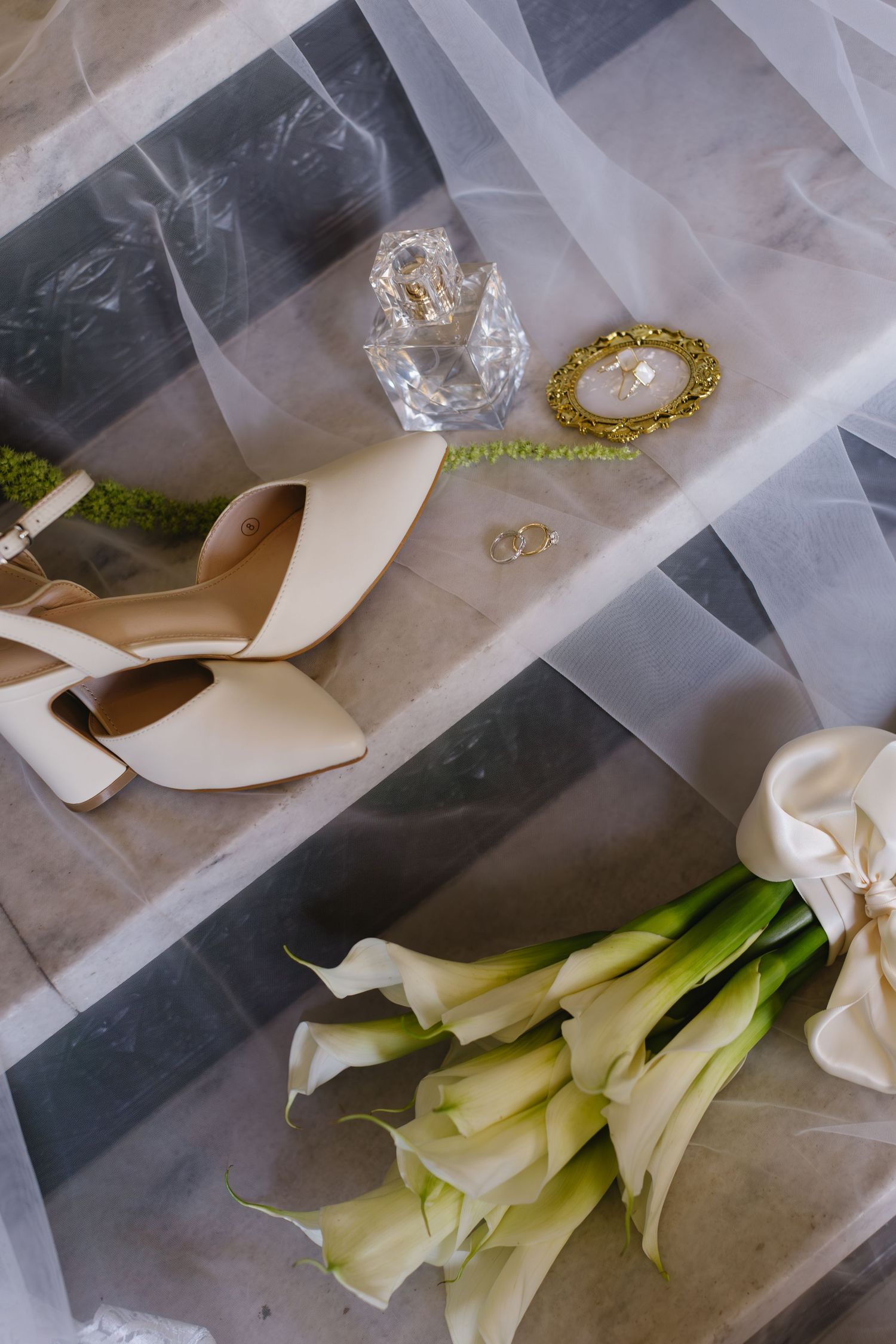 Wedding accessories including white shoes, jewelry, and calla lily bouquet arranged on gray marble surface.