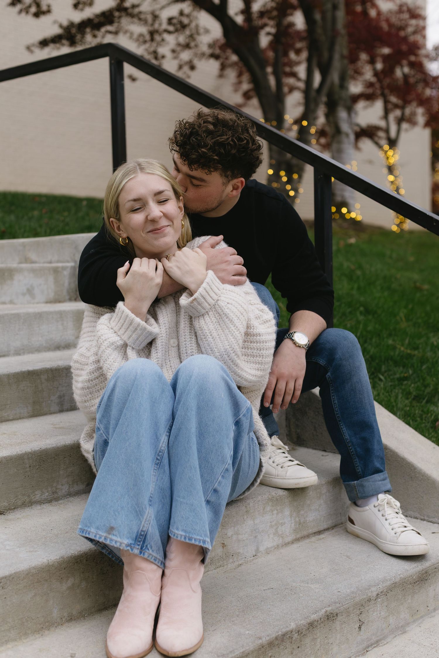 A couple sits together on concrete steps, wearing casual attire including jeans and sweaters on an autumn day.
