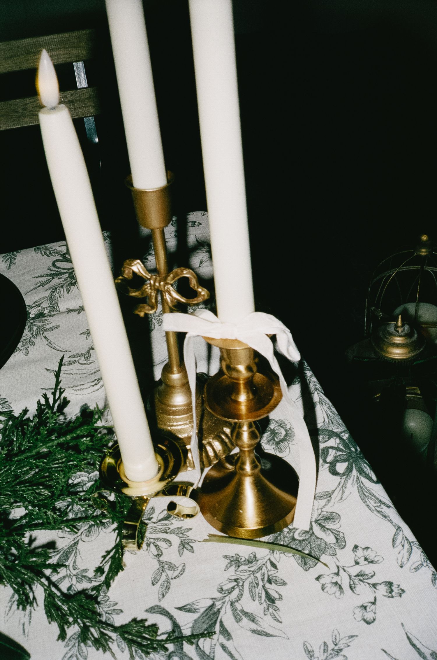 White taper candles in brass holders on a table with greenery and floral print linens.