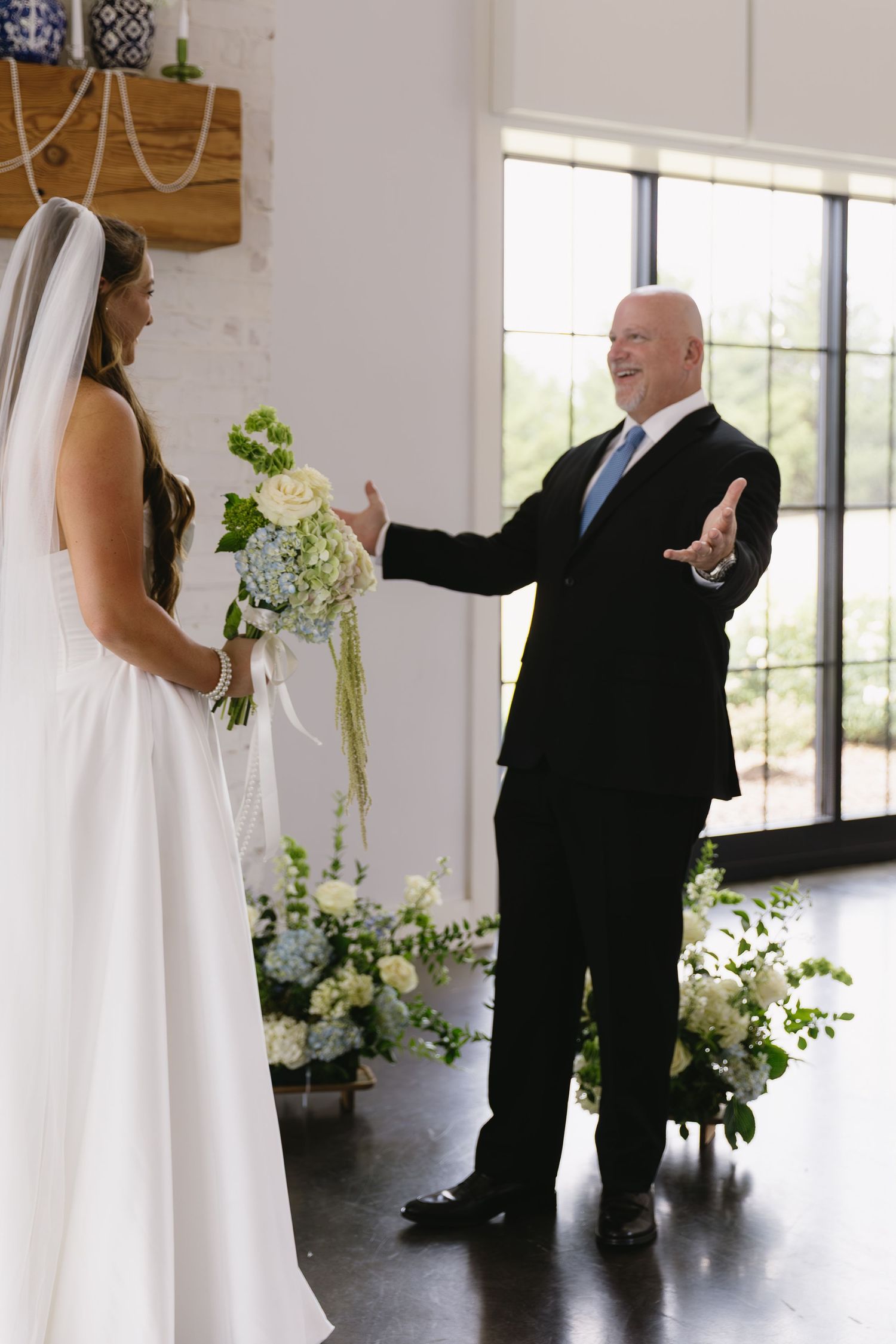 First look wedding moment in a bright indoor venue with white walls and floral decorations.
