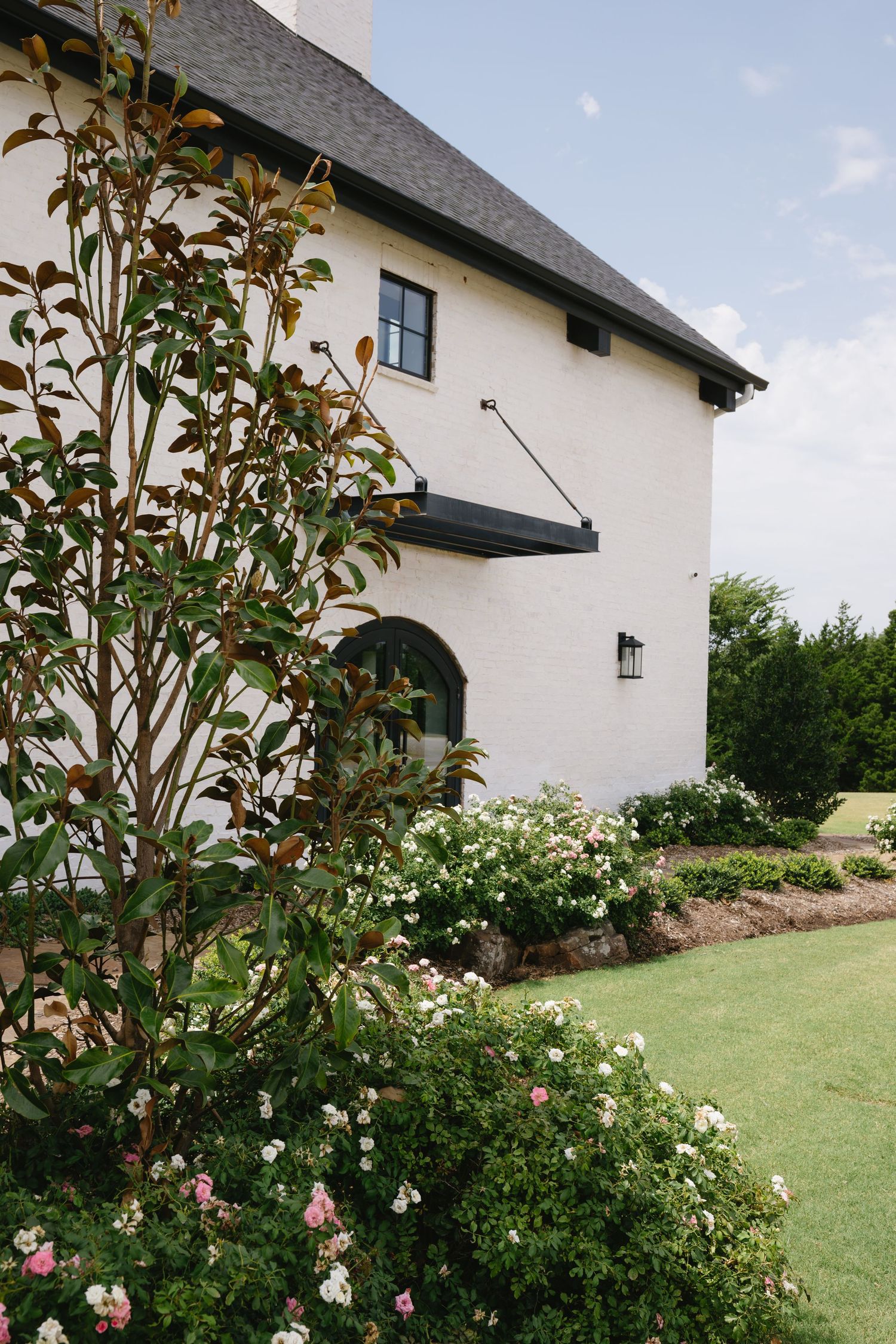White house exterior with lush landscaping and flowering bushes against a blue sky.