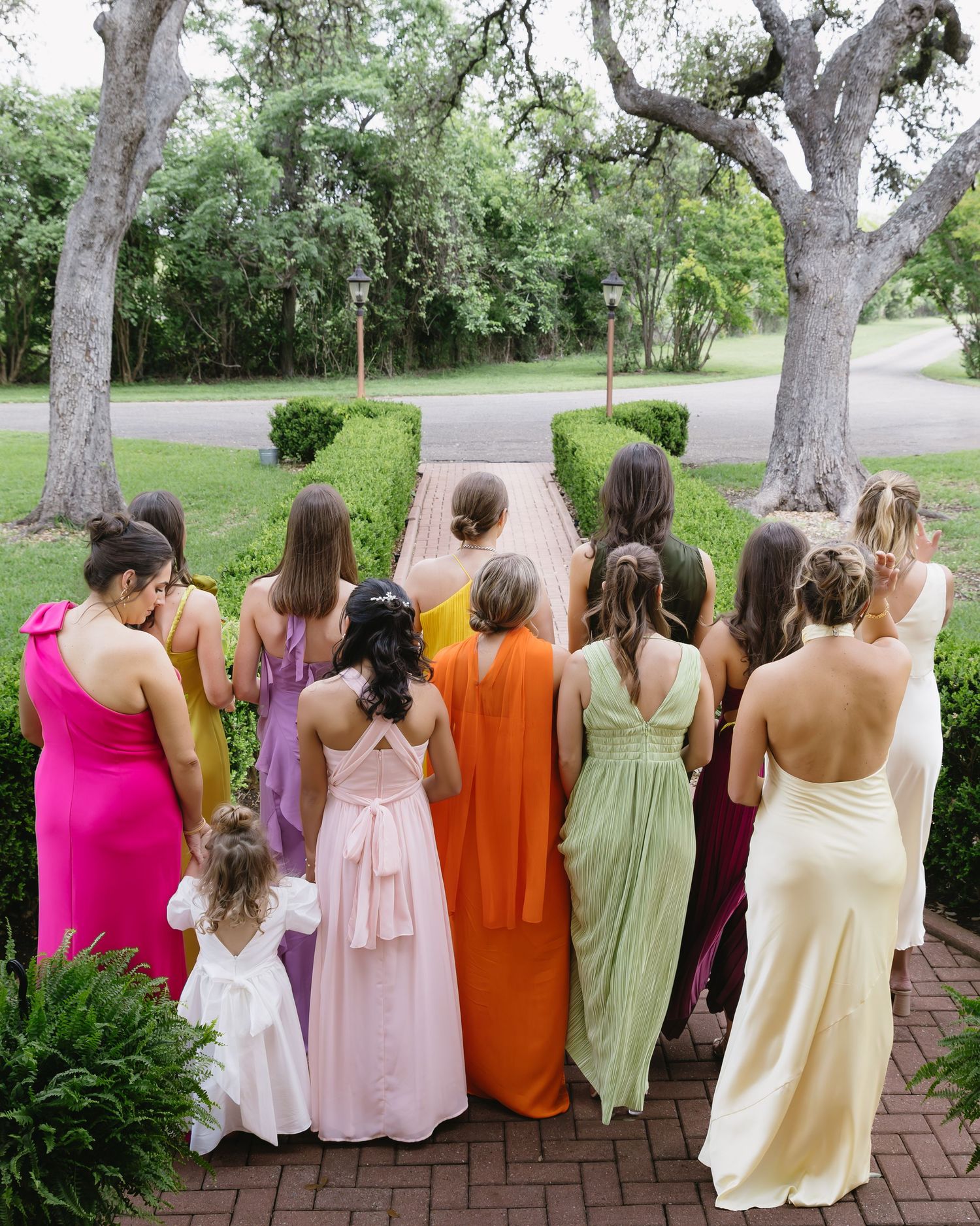 A group of bridesmaids in colorful dresses standing together on a brick pathway surrounded by trees.