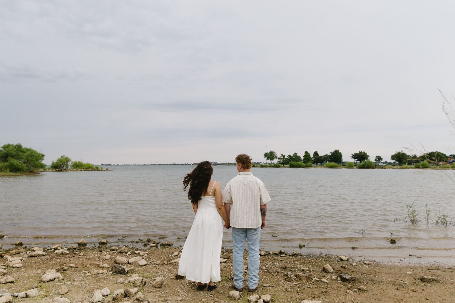 Wide landscape shot of couple standing by lake shore during cloudy day.