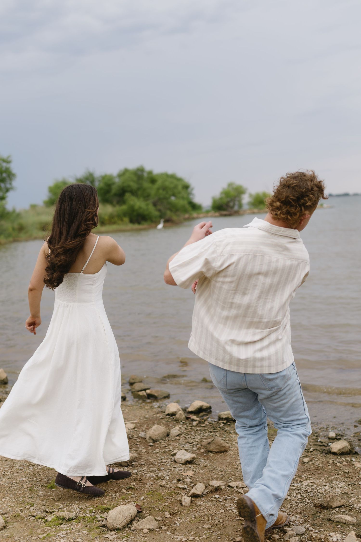 Two people in white outfits dance playfully by the water's edge.