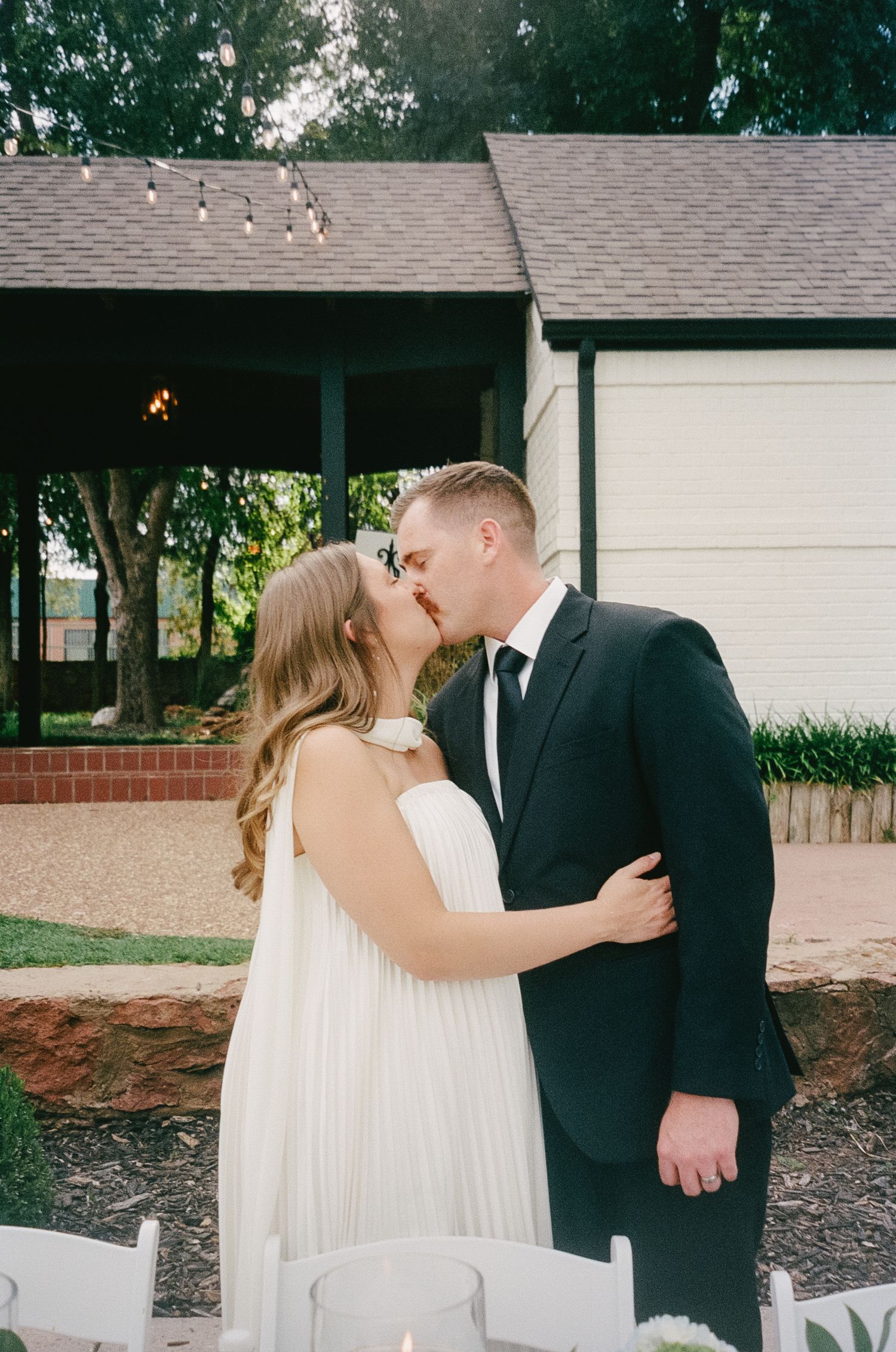 Romantic outdoor wedding moment captured against white building with string lights overhead.