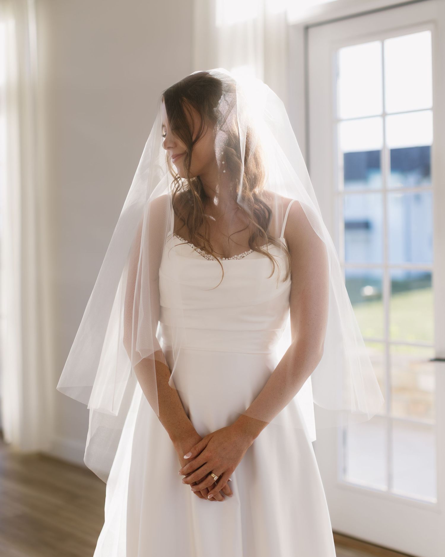 A bride in a sleeveless white wedding gown stands by bright windows in a contemporary indoor space.