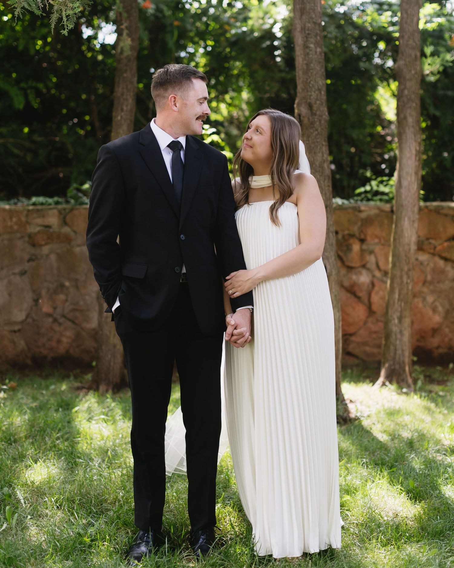 Wedding couple shares intimate moments against stone wall and trees in outdoor garden setting.